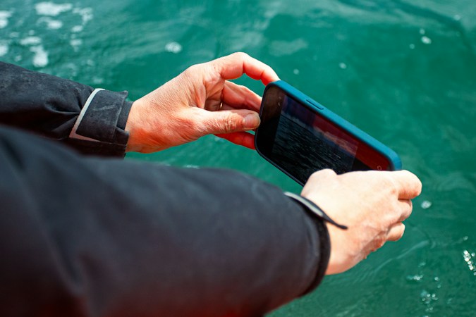 Two hands in a dark jacket sleeve holding a phone in a blue case sideways to take a photo over clear green seawater