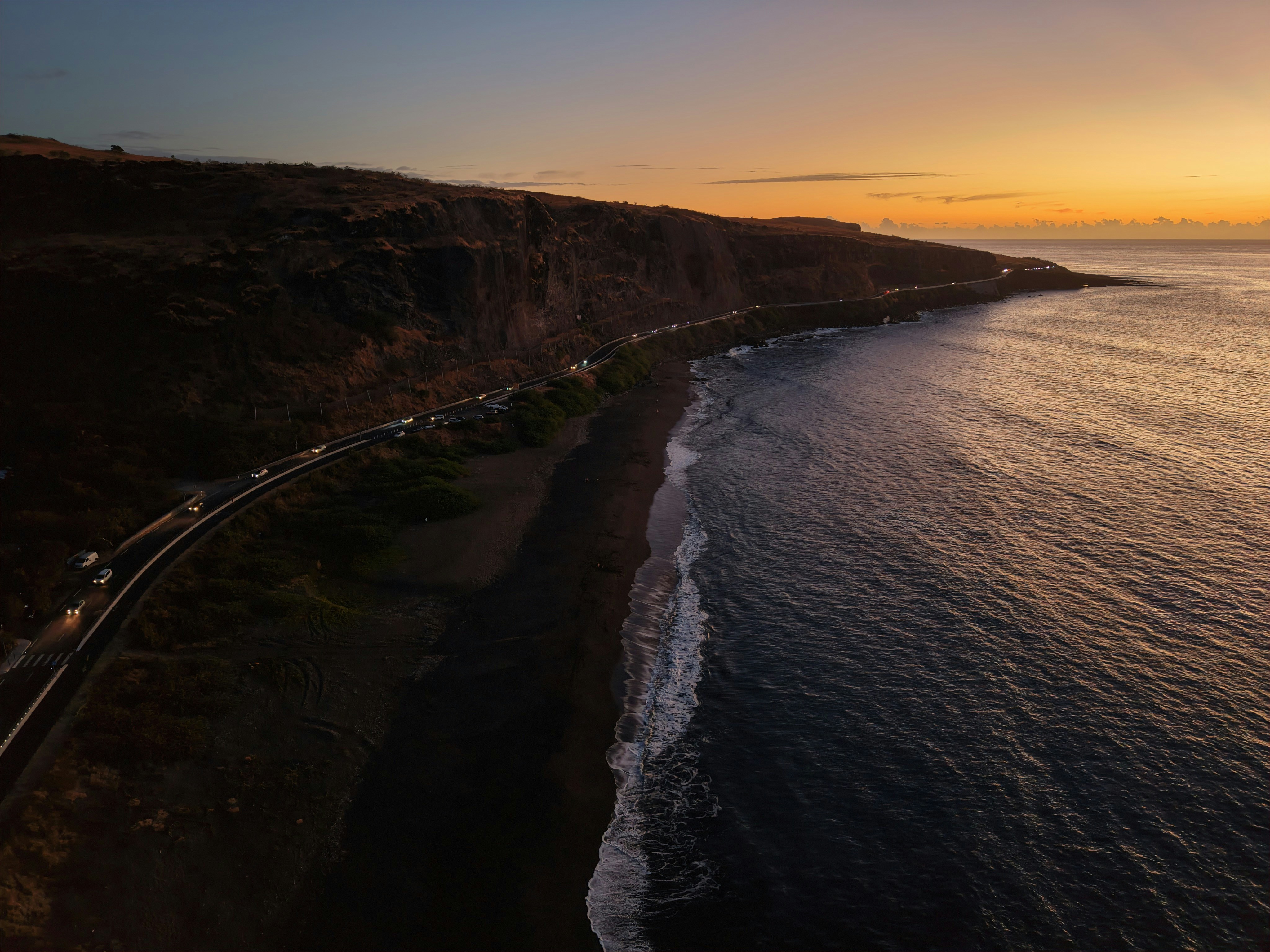 Coastal road alongside a dark beach and ocean at sunset.