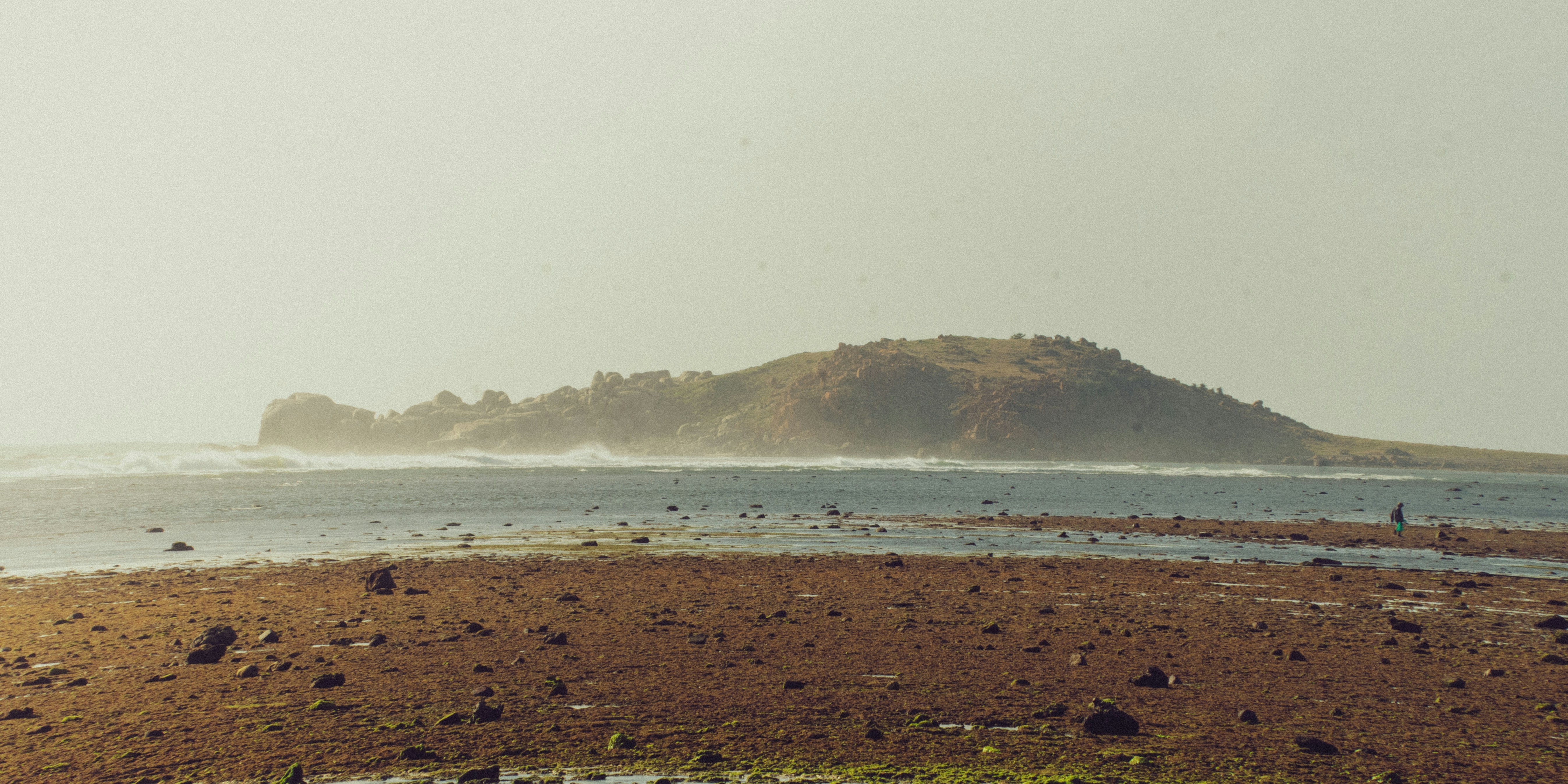 Felsiger Strand mit entfernter Insel und dunstigem Himmel.
