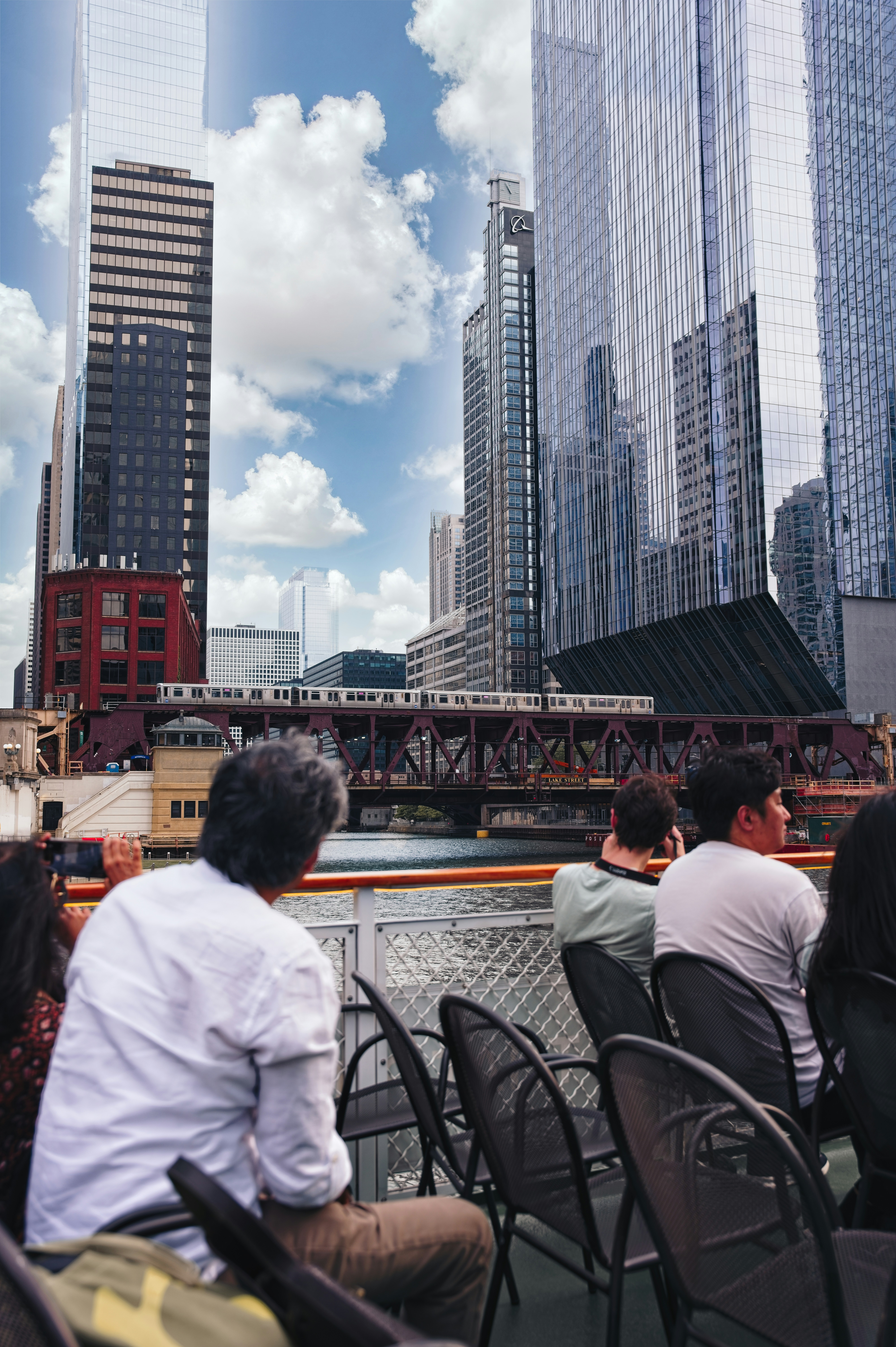 People seated along a riverfront with a backdrop of tall glass skyscrapers and a red bridge, capturing a cityscape from a ferry deck. This is a candid urban scene.
