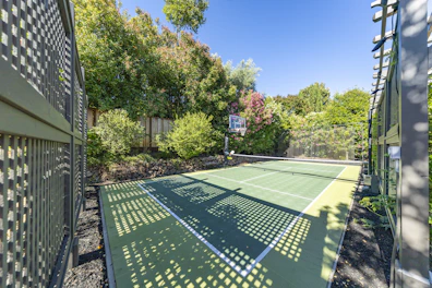Outdoor pickleball court with basketball hoop surrounded by lush greenery.