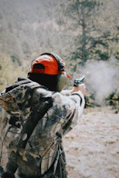 Person in camouflage firing a handgun outdoors with muzzle flash.