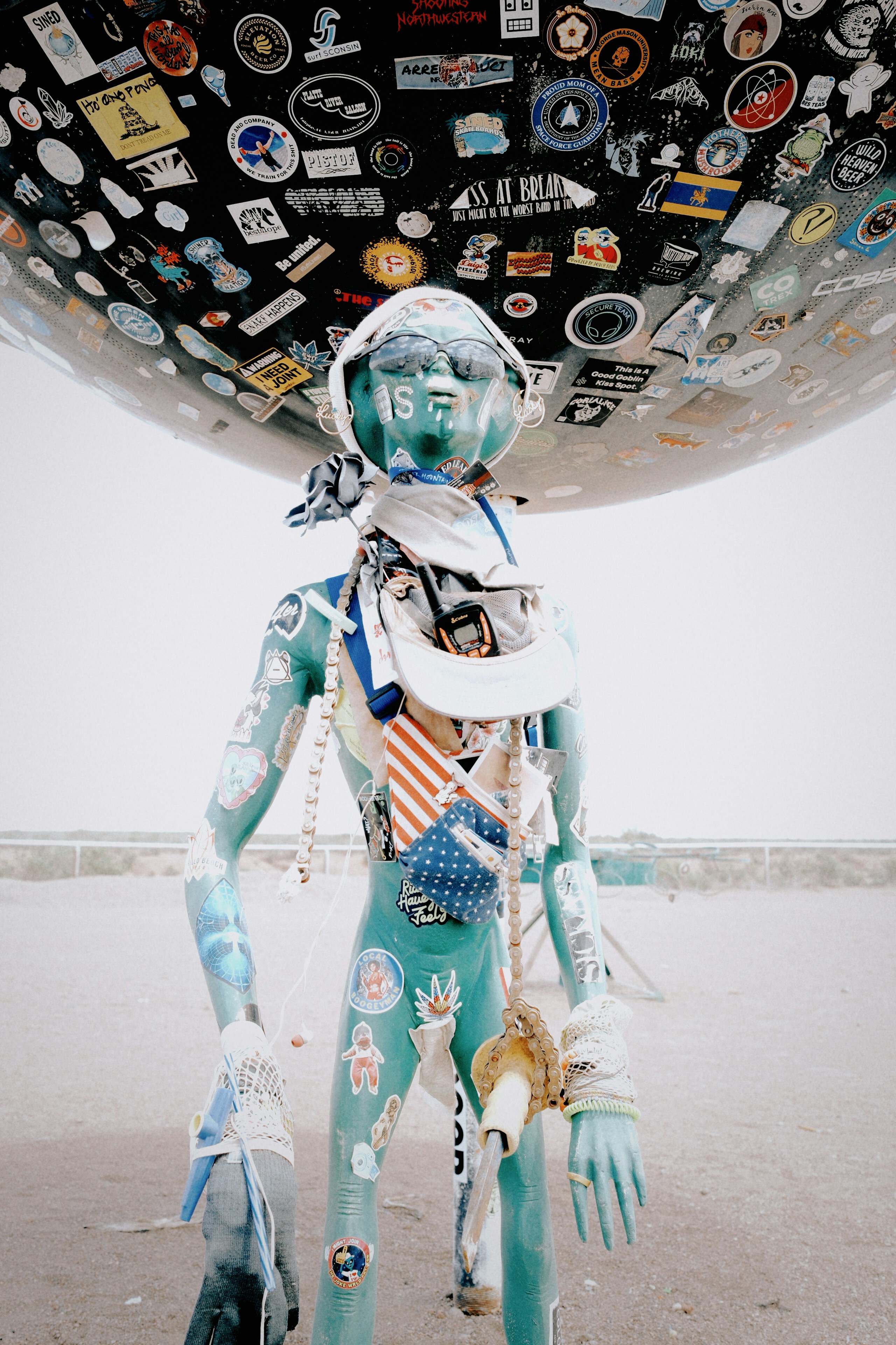 A turquoise mannequin with stickers under a large sticker-covered sphere.