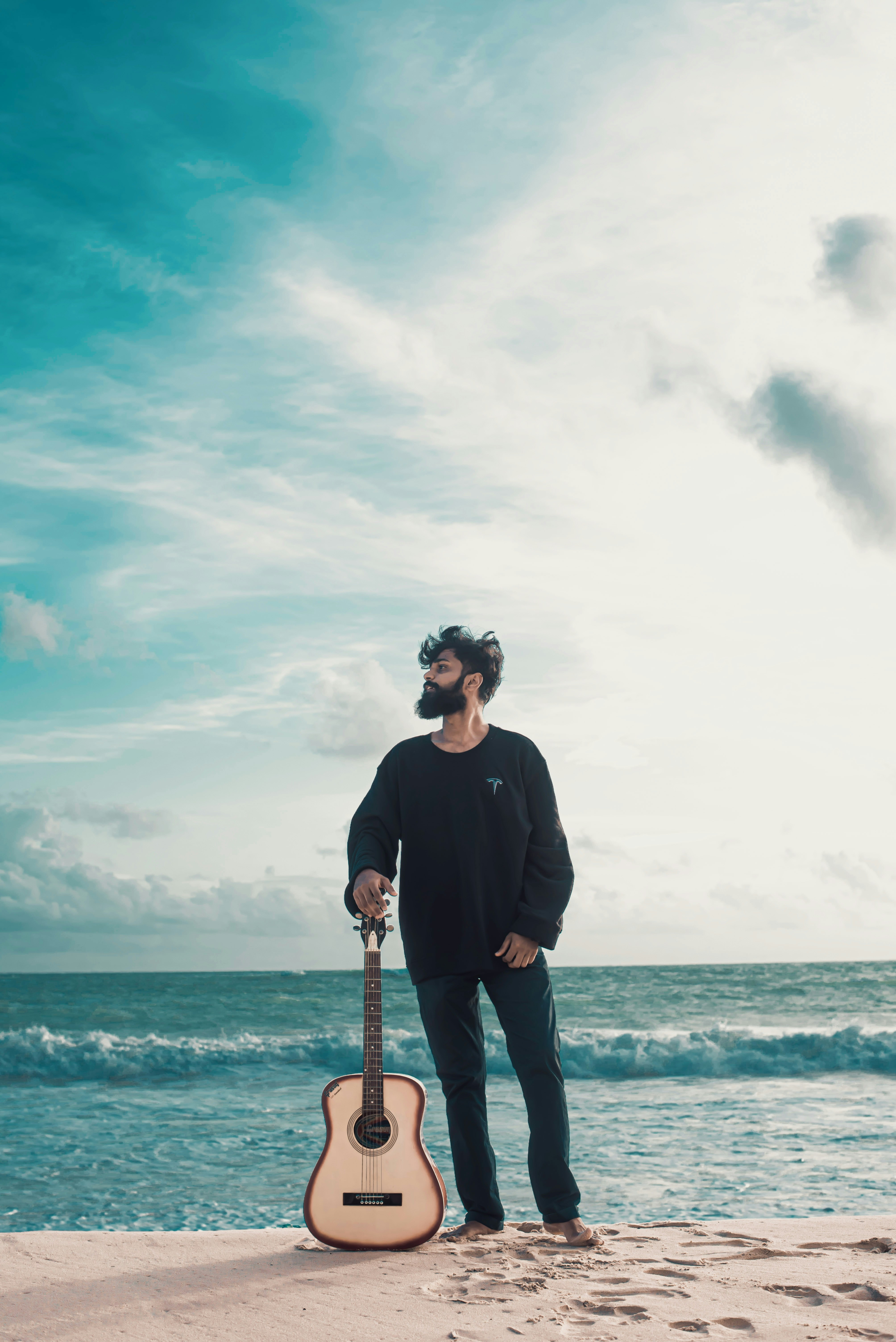 Man with guitar stands on sandy beach by the ocean.