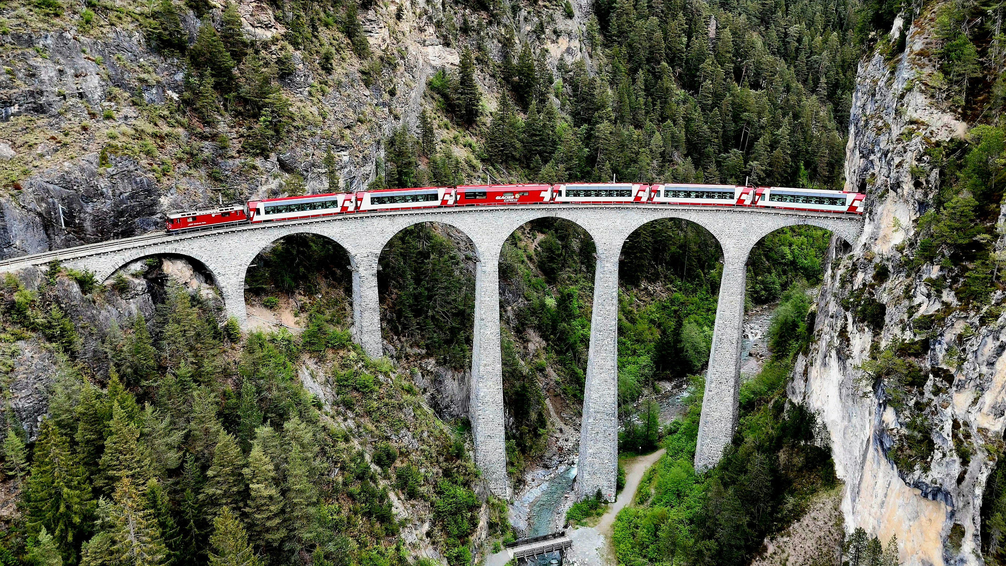 Trem vermelho cruzando um viaduto alto em um vale arborizado.