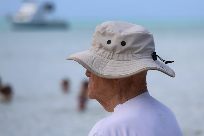 Older man in a stone-colored bucket hat with a chin cord and toggle, seen in profile against a blurred turquoise sea