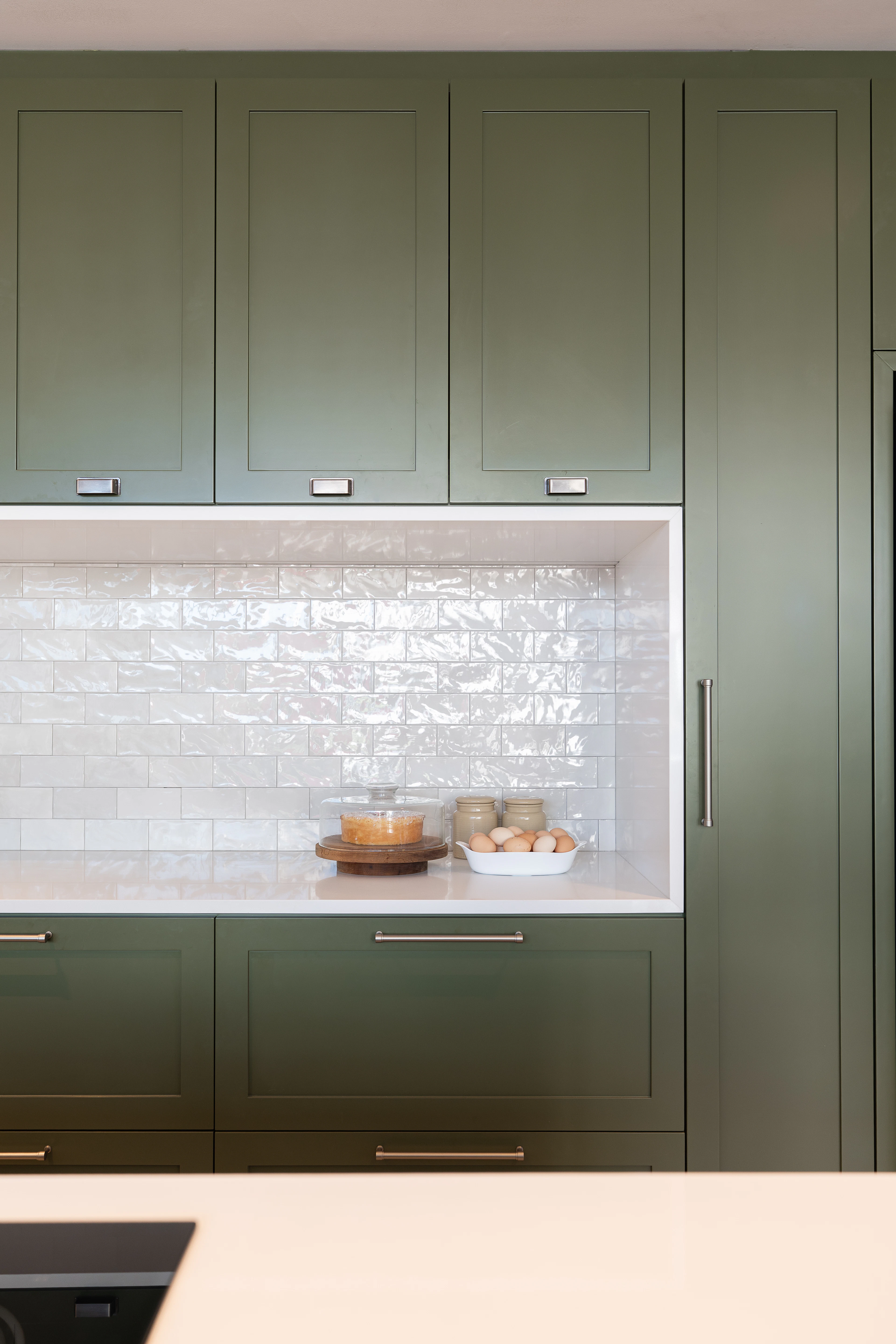 Sage-green kitchen cabinets around a white tiled niche, with a clear countertop