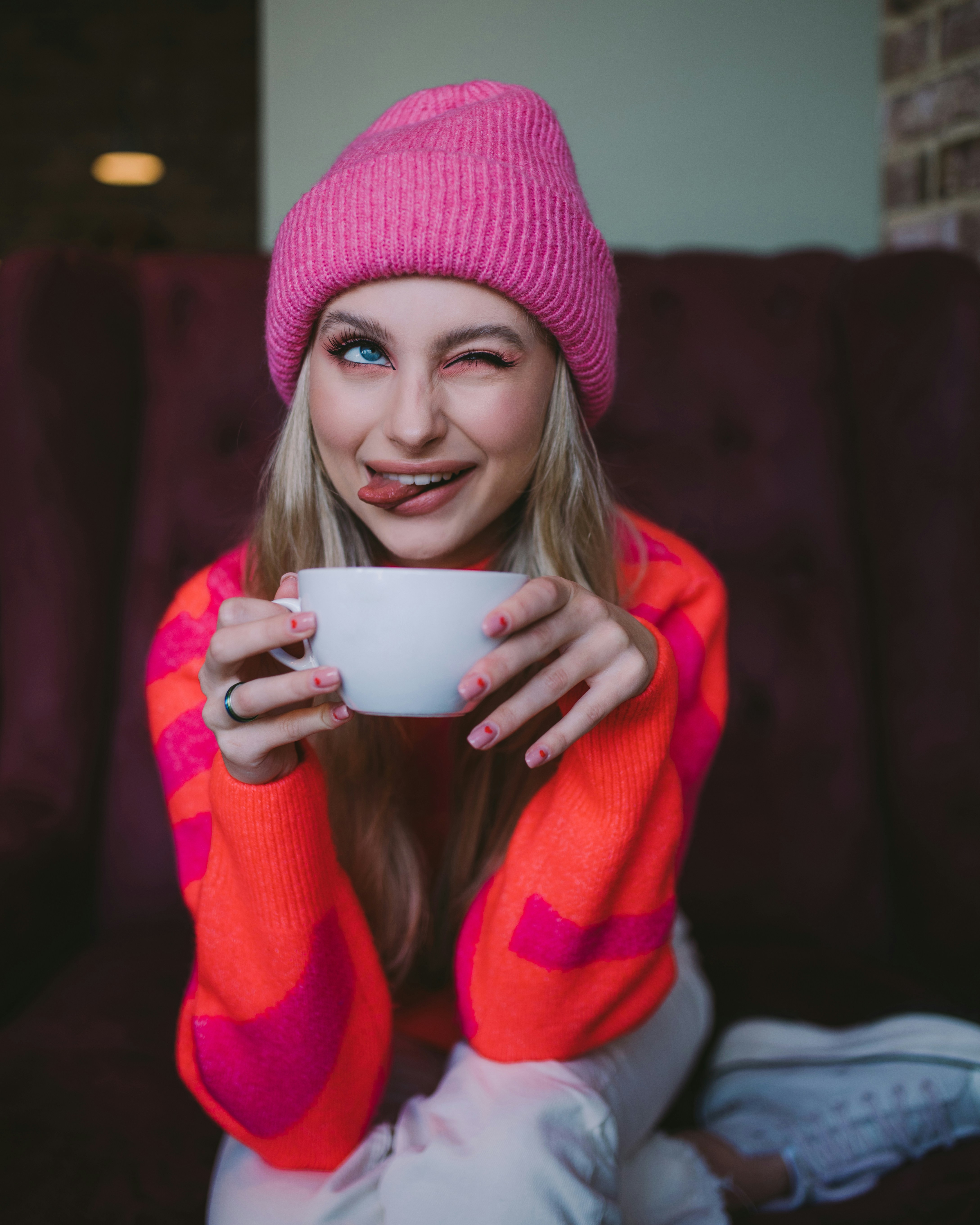 Young woman in pink beanie winks, holding coffee cup.