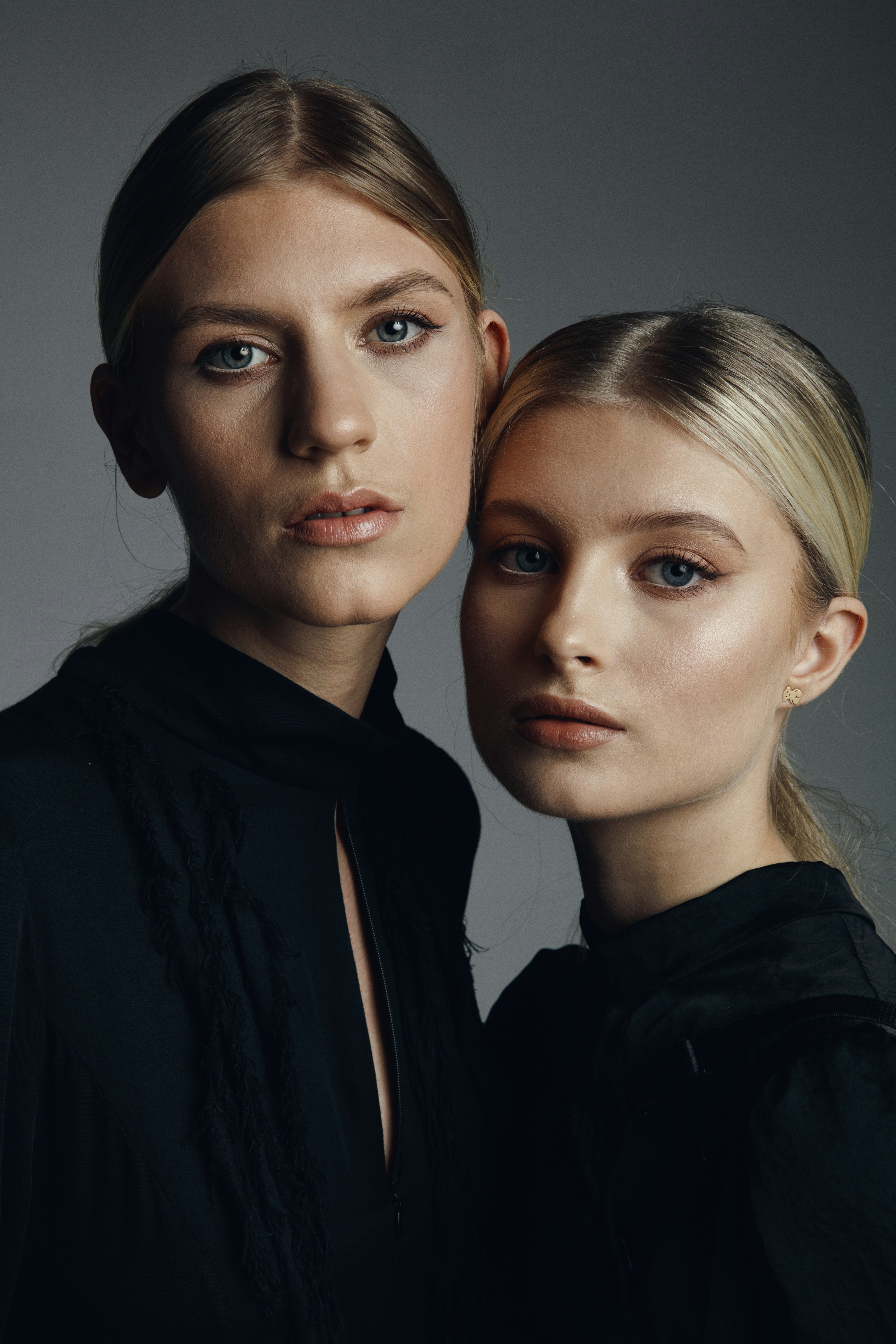 Two blonde women in black tops against a grey background.