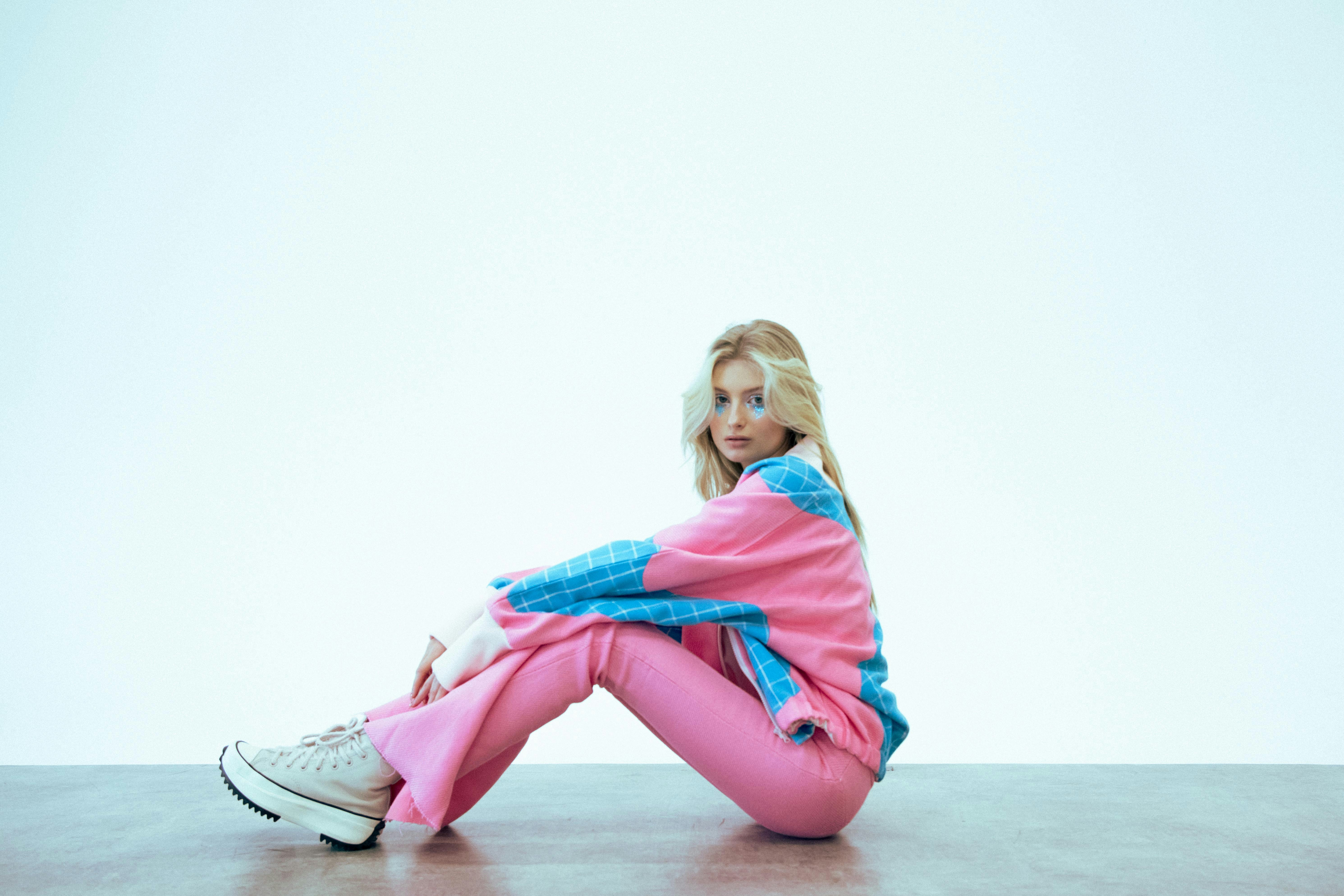 Blonde woman in pink and blue tracksuit sitting on floor.