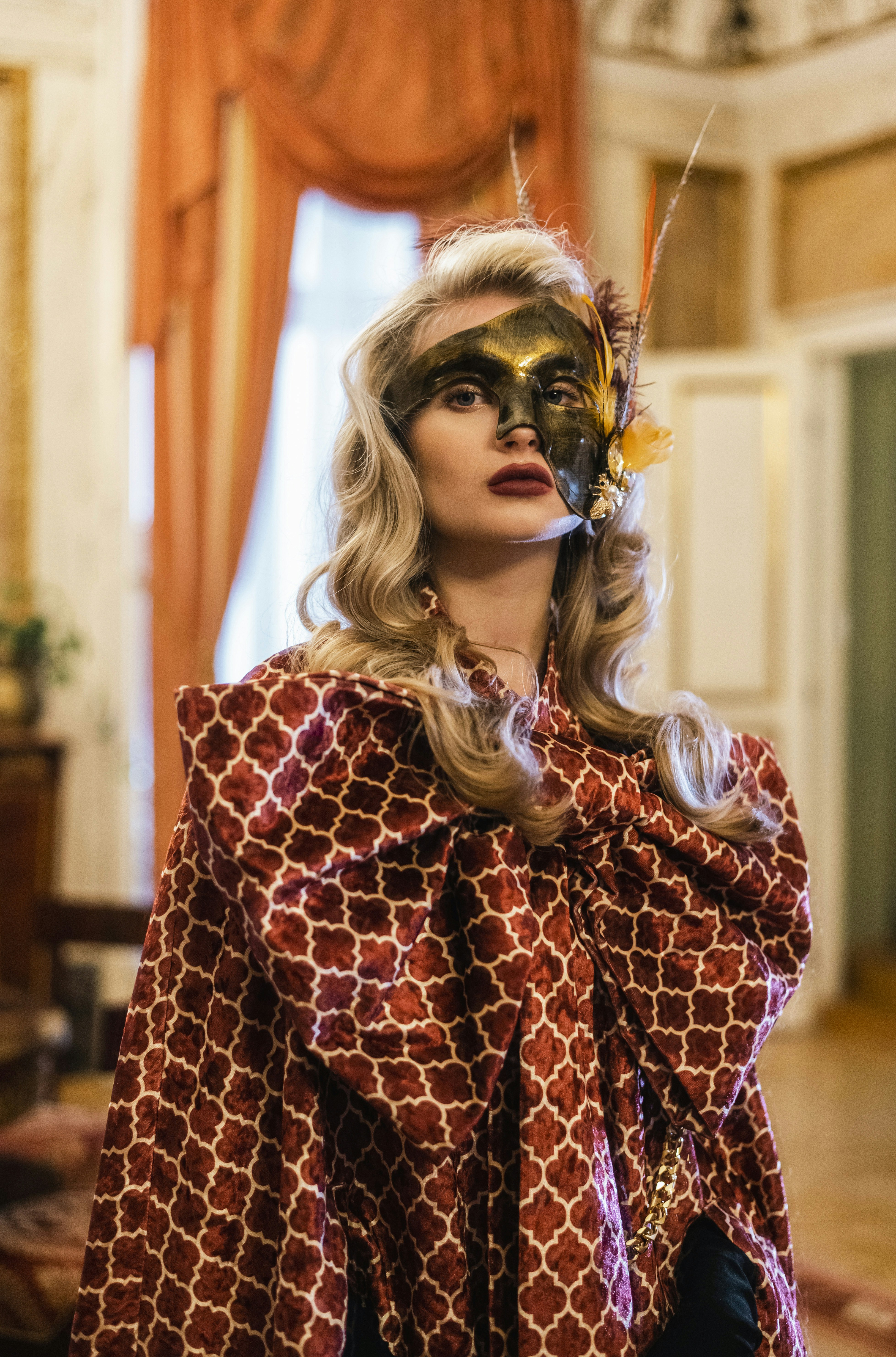 Wanita elegan mengenakan topeng dekoratif dan jubah bermotif di dalam ruangan.