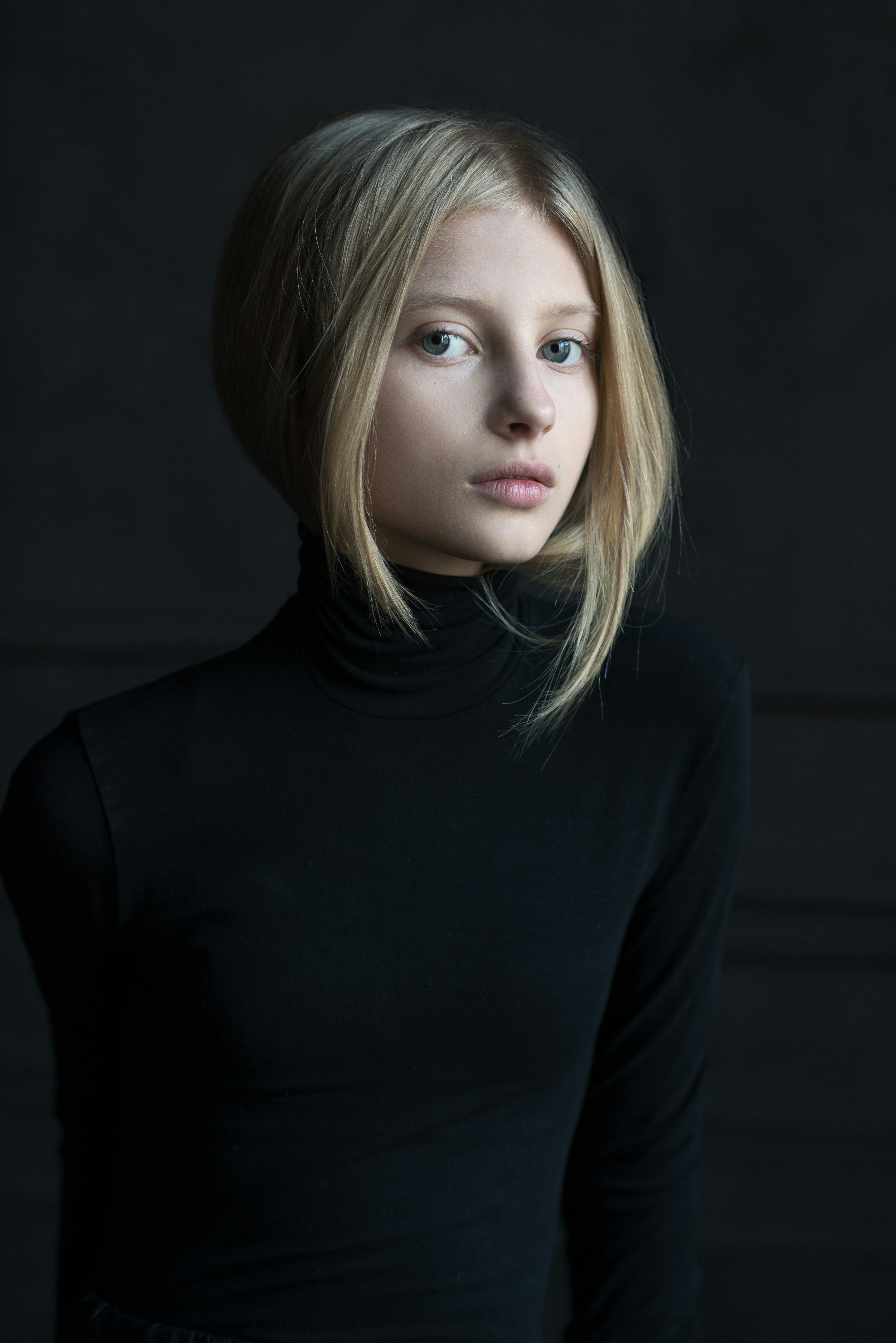 Blonde woman in black turtleneck against dark background.