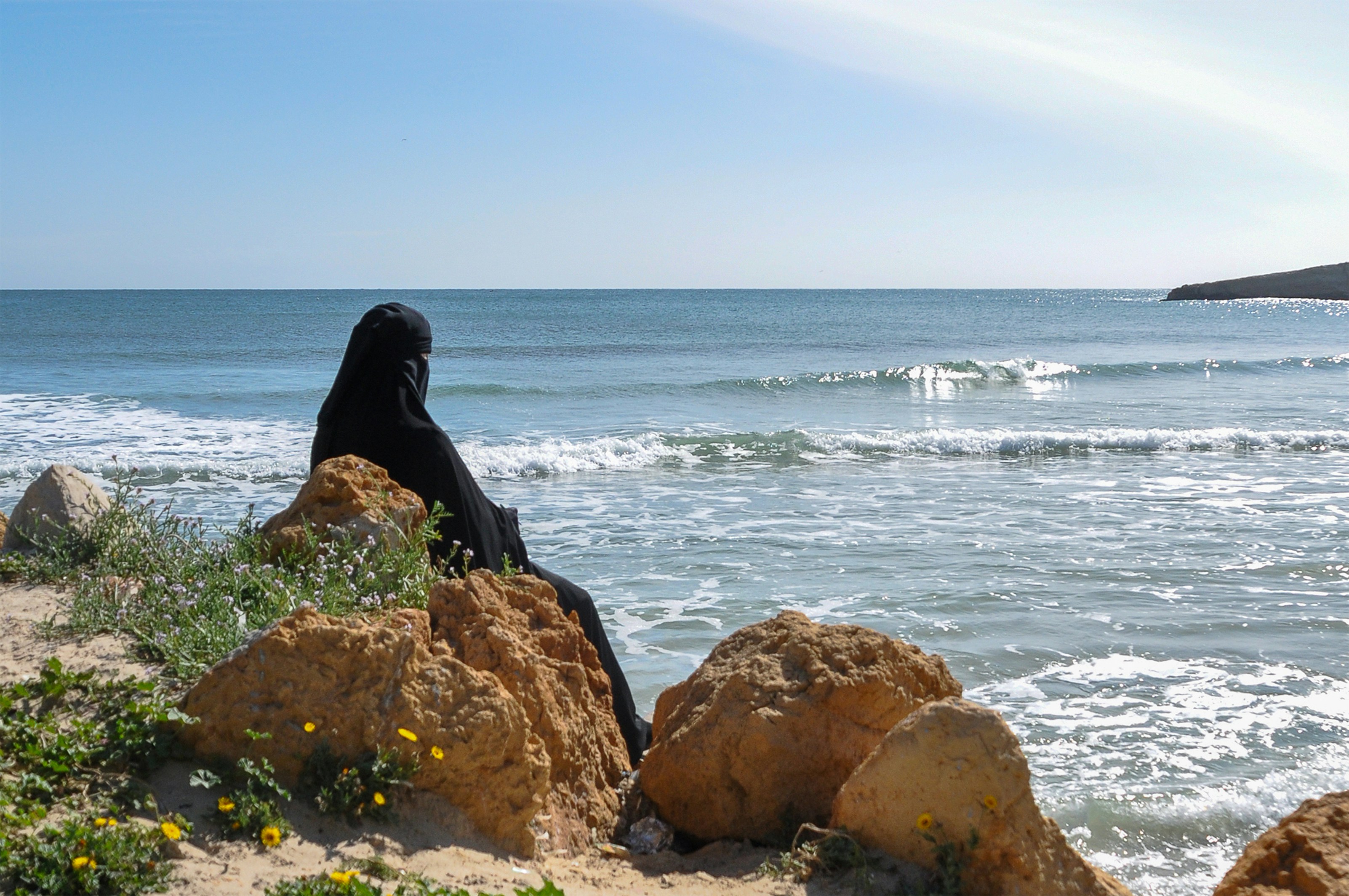 Mujer con niqab sentada en rocas junto al mar.