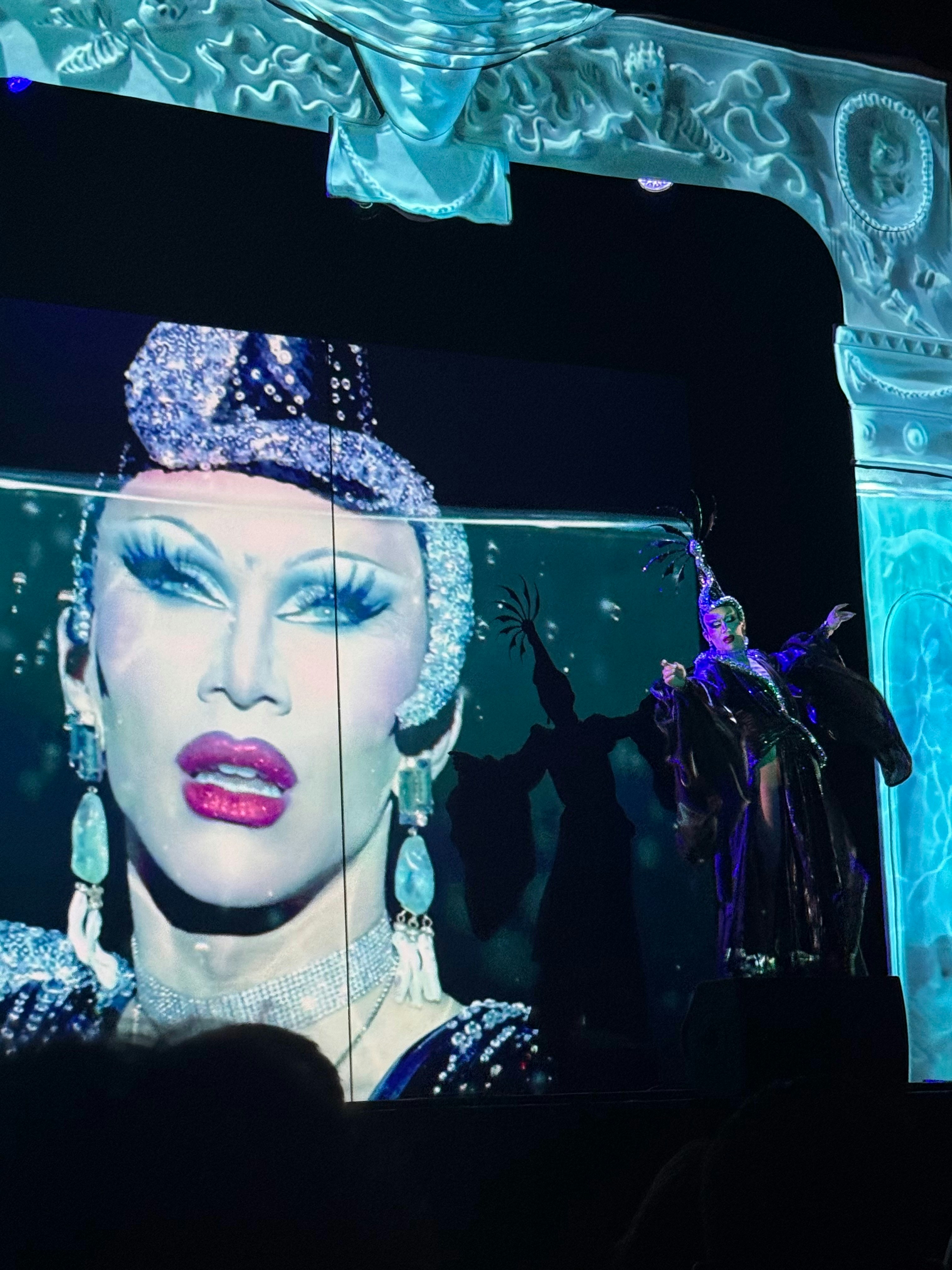 Drag queen performing on stage with a large screen display.