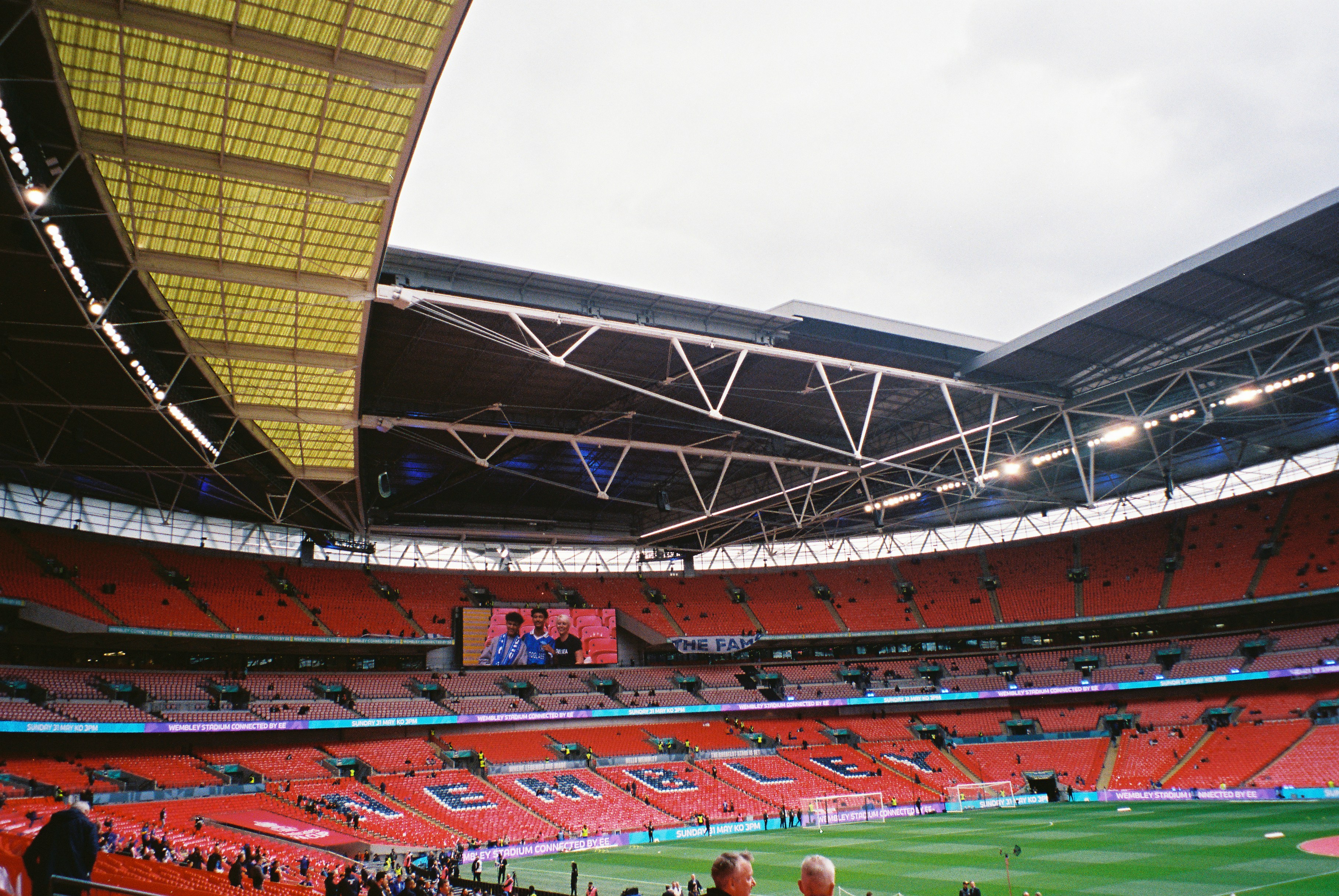 Stadion kosong dengan kursi merah dan layar besar.