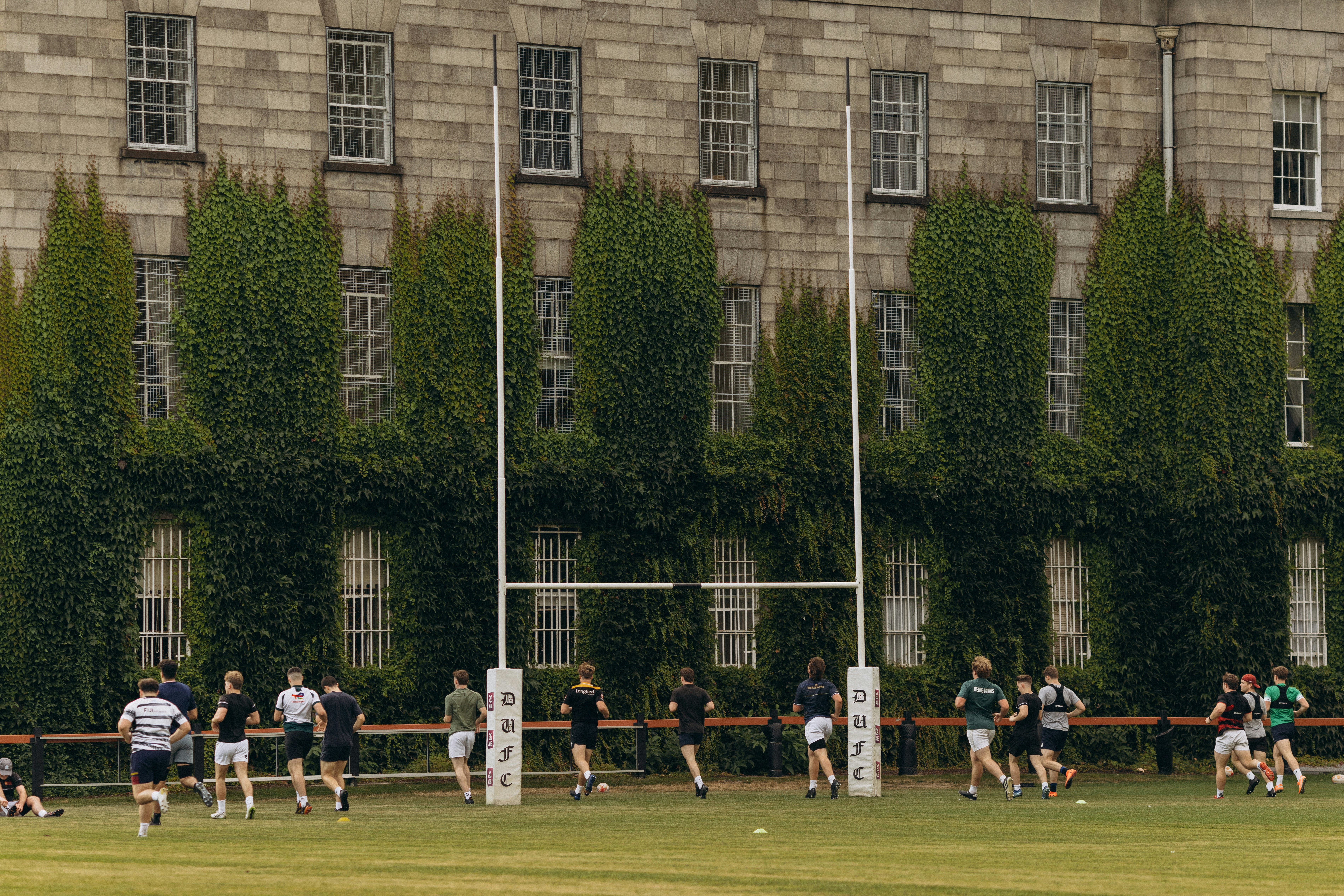 Rugby players practice near goalposts with an ivy-covered building backdrop.