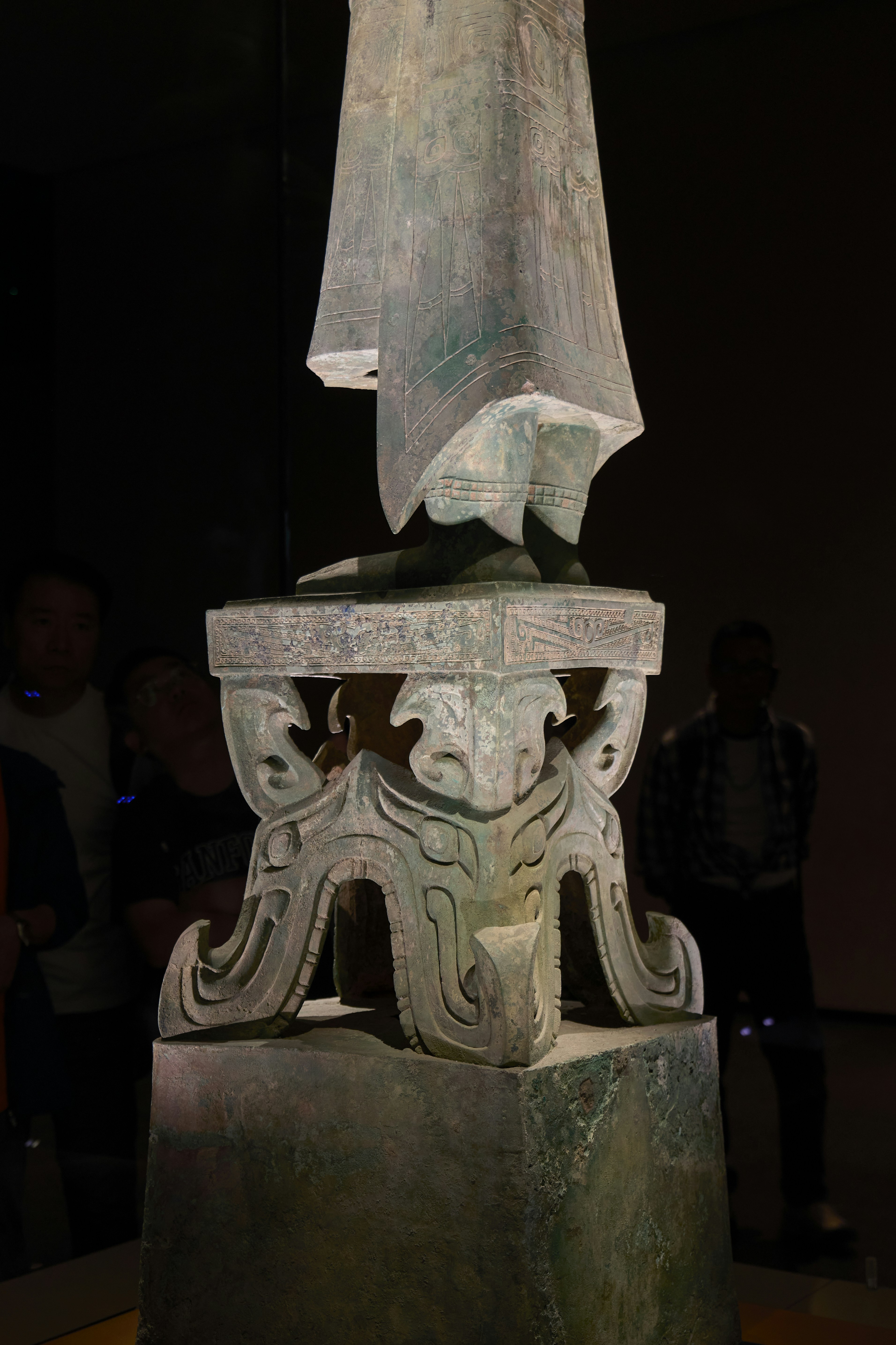 Base de estátua antiga de bronze com entalhes intrincados sobre fundo escuro.