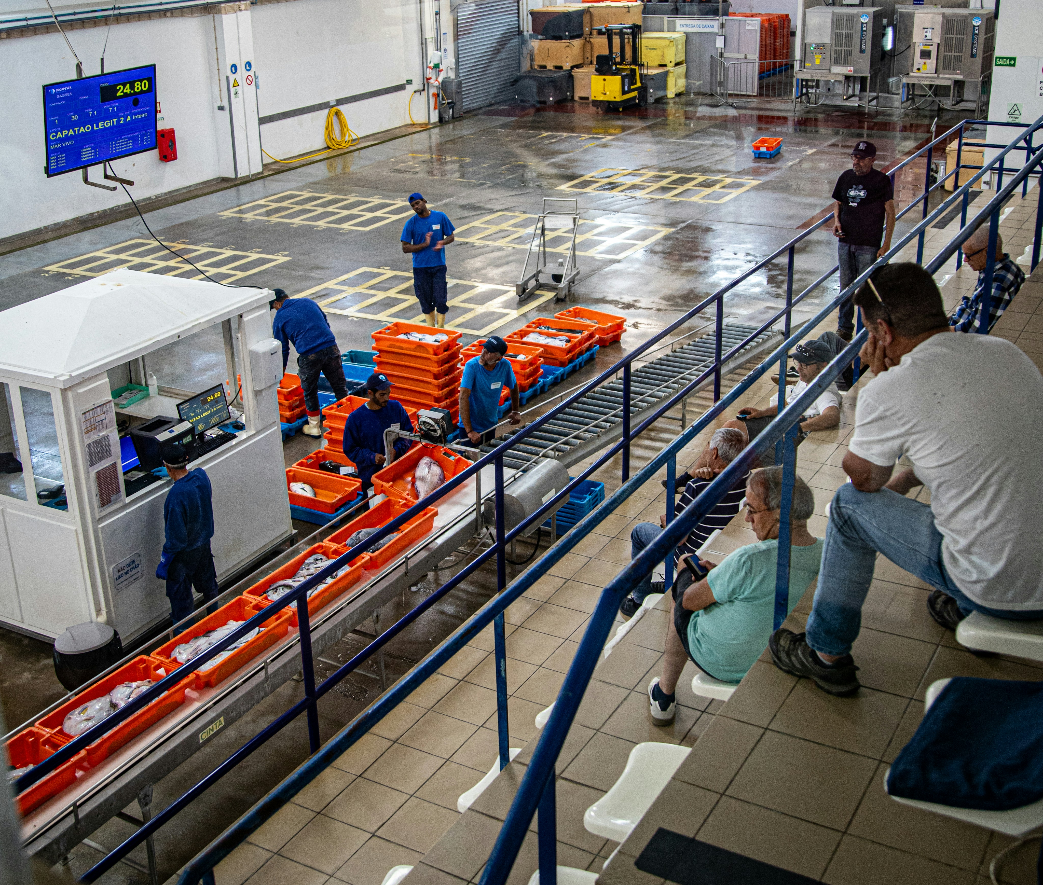 Industrial facility with workers moving crates and observers watching.