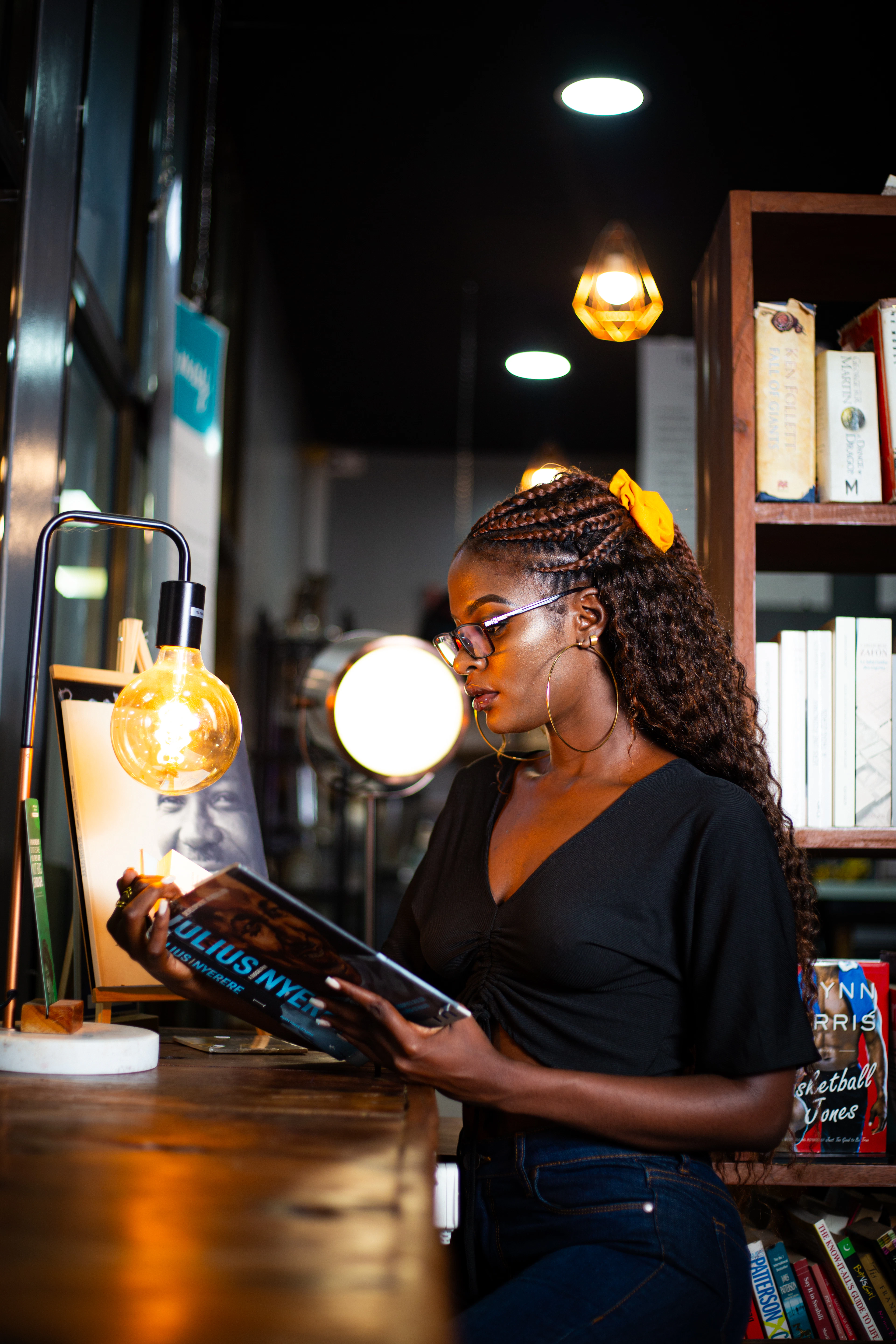 A woman with a yellow hair clip reading a book in a warmly lit bookstore, bulb lights glowing behind her