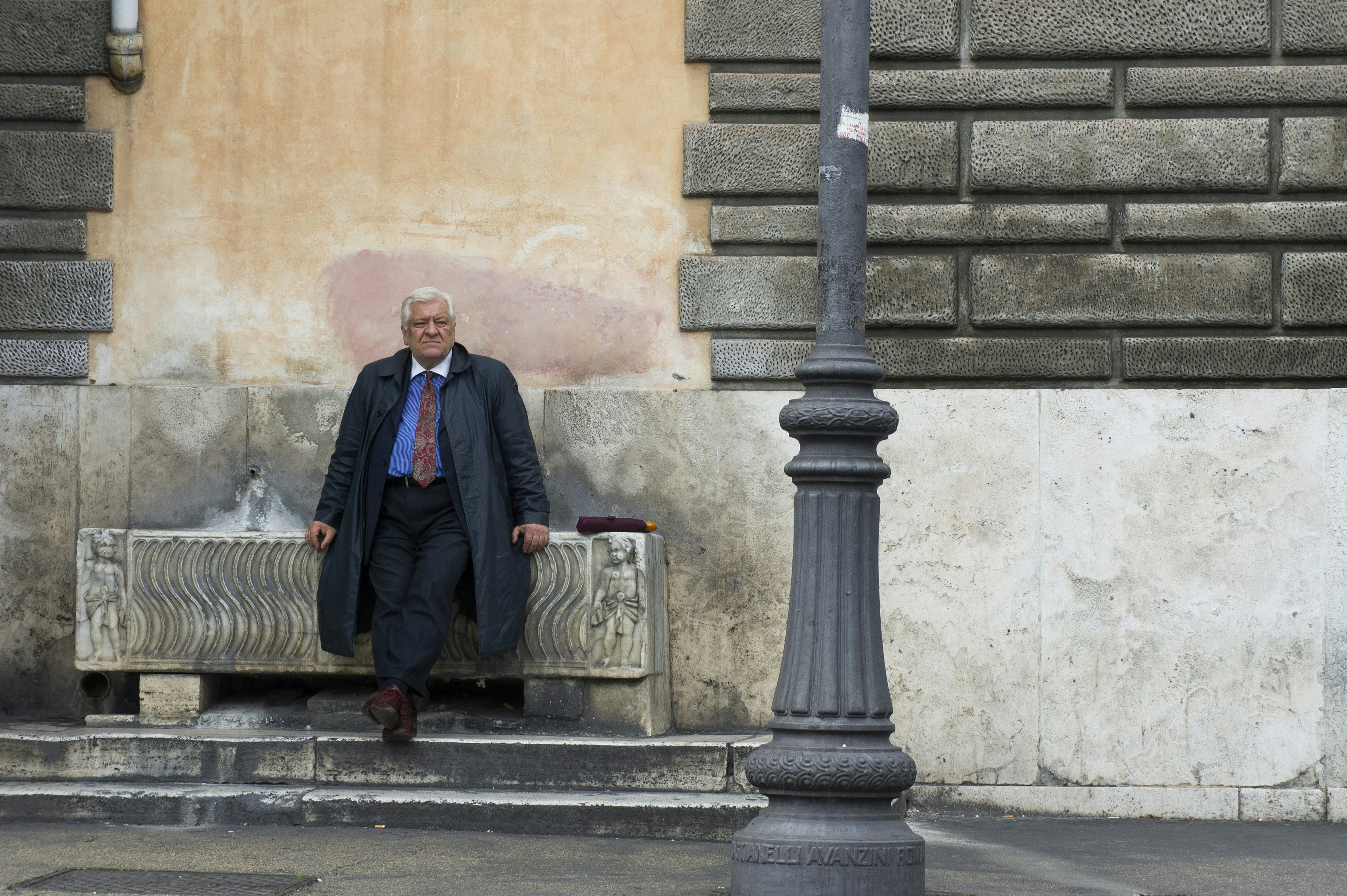 A man in a dark coat sits by a lamppost.