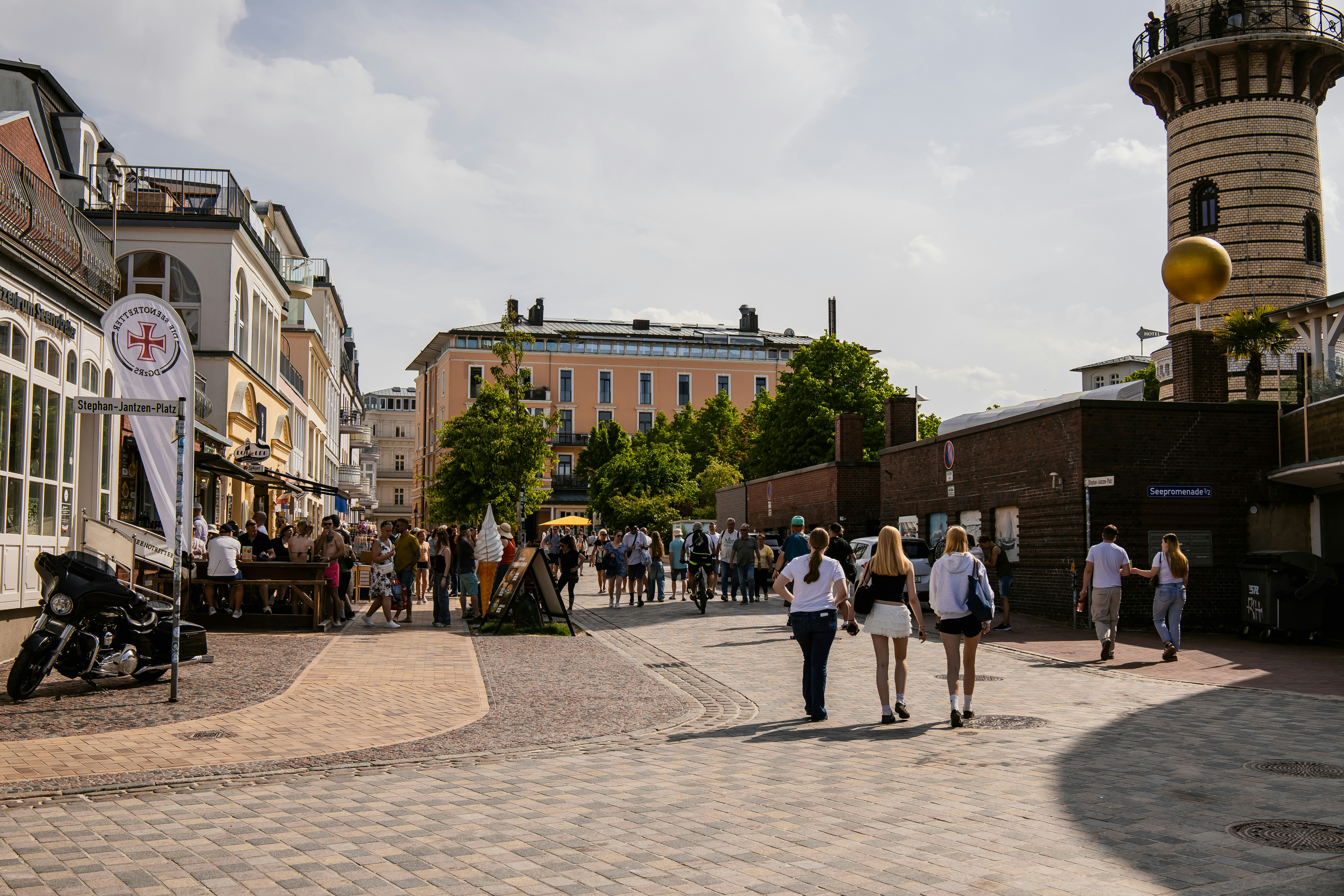 Sunny street with people, shops, and a distinctive tower.