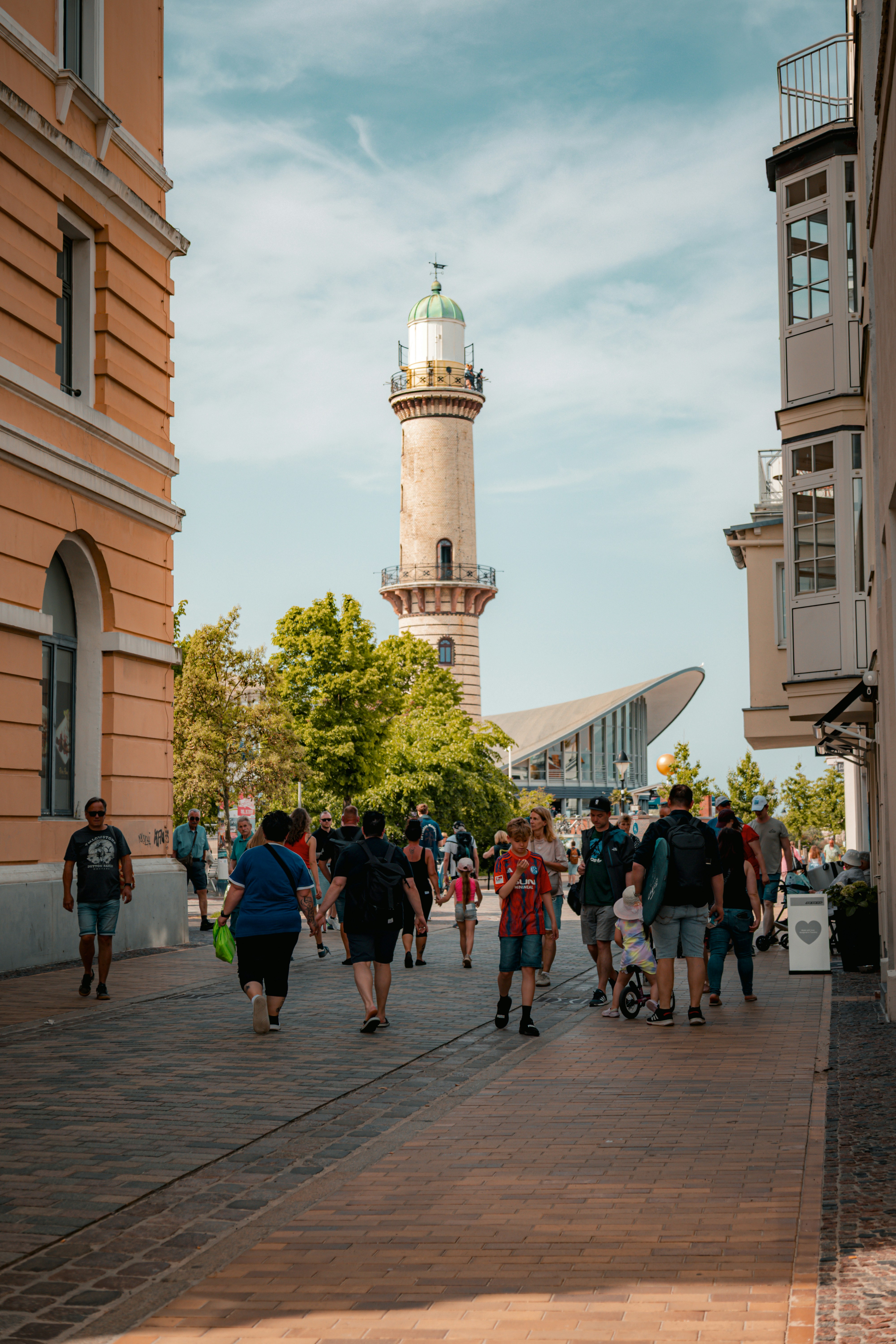 People walk down a street towards a lighthouse and buildings.