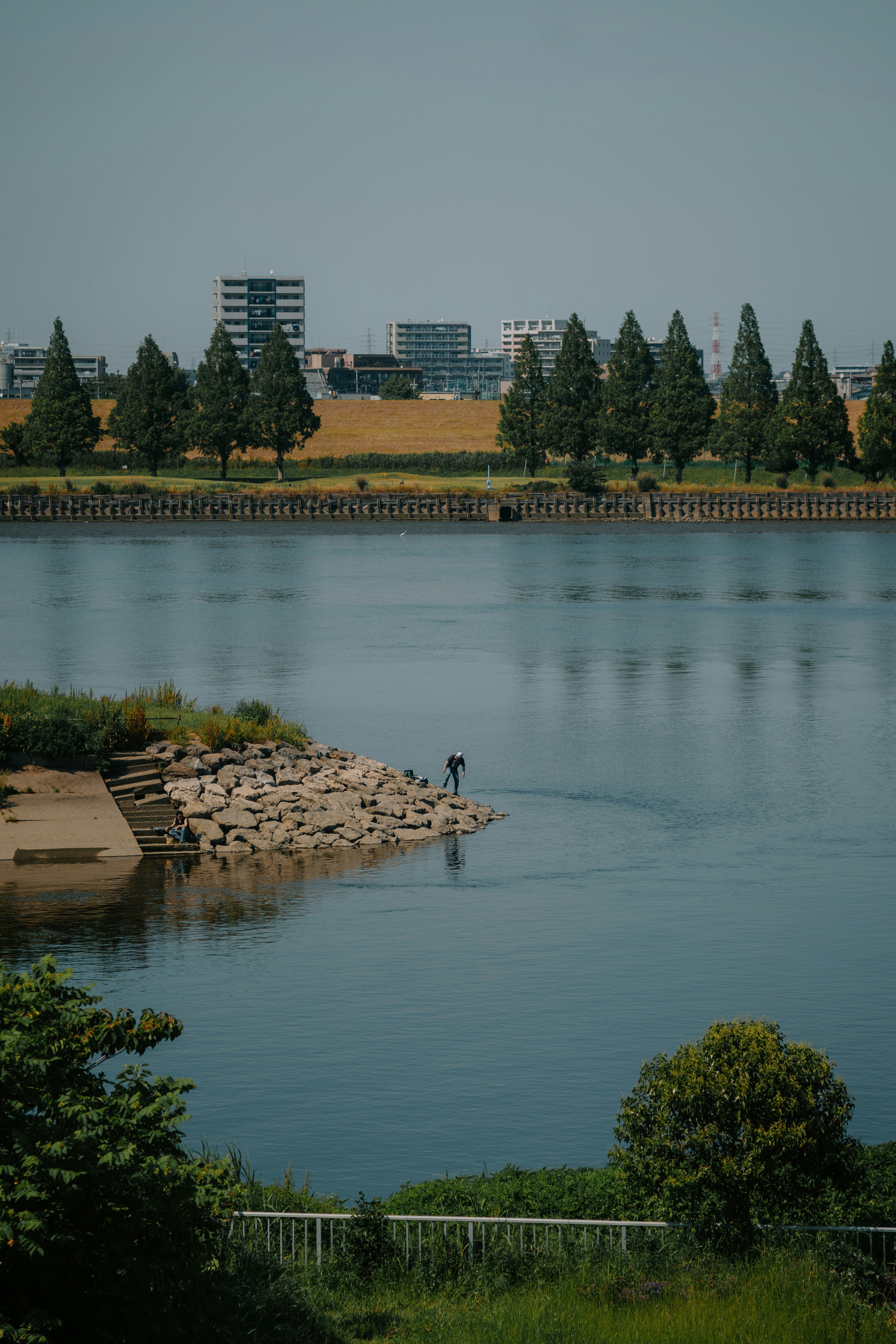 A person stands on a rocky riverbank with city buildings.