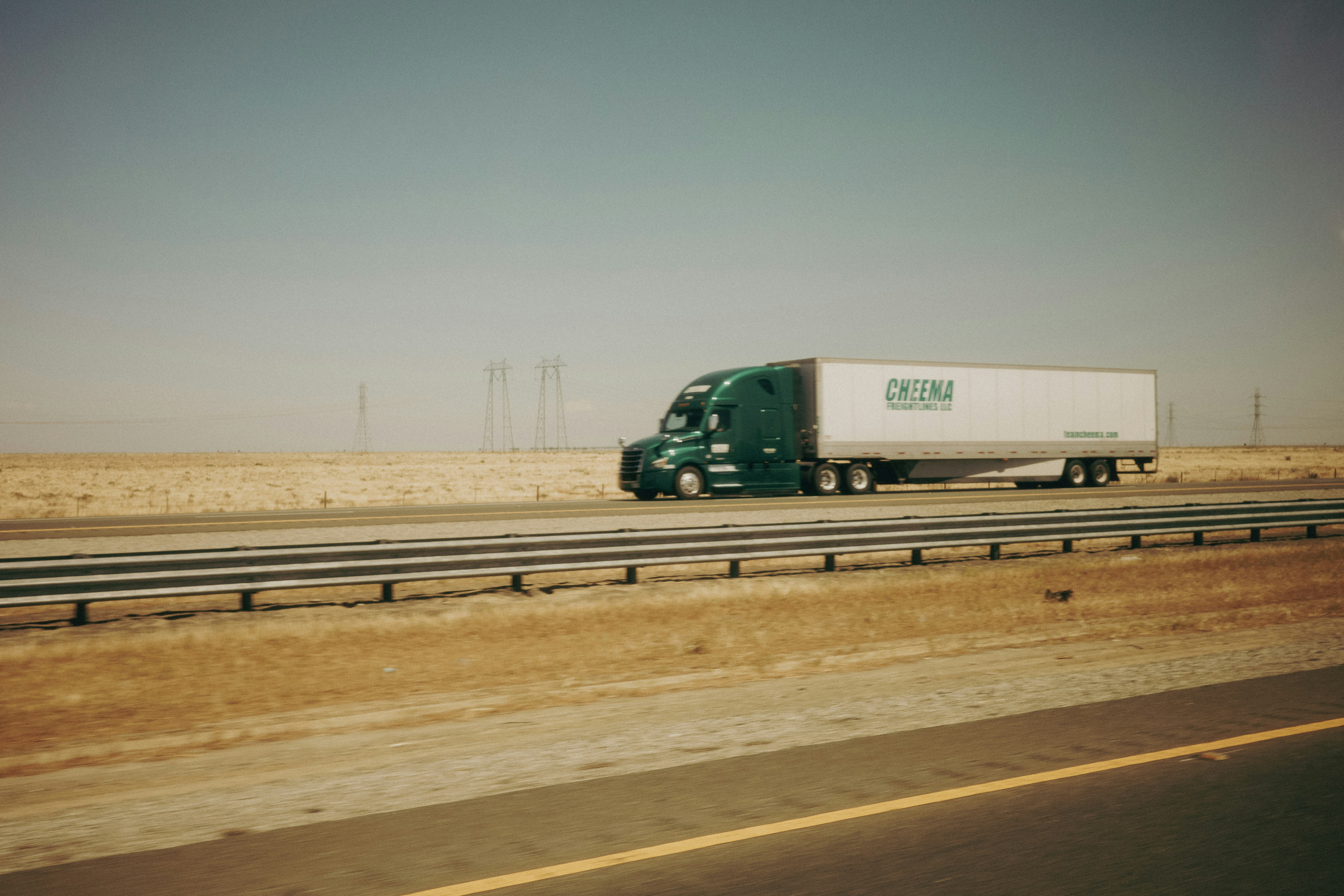 Green semi-truck on highway with dry landscape and wind turbines.