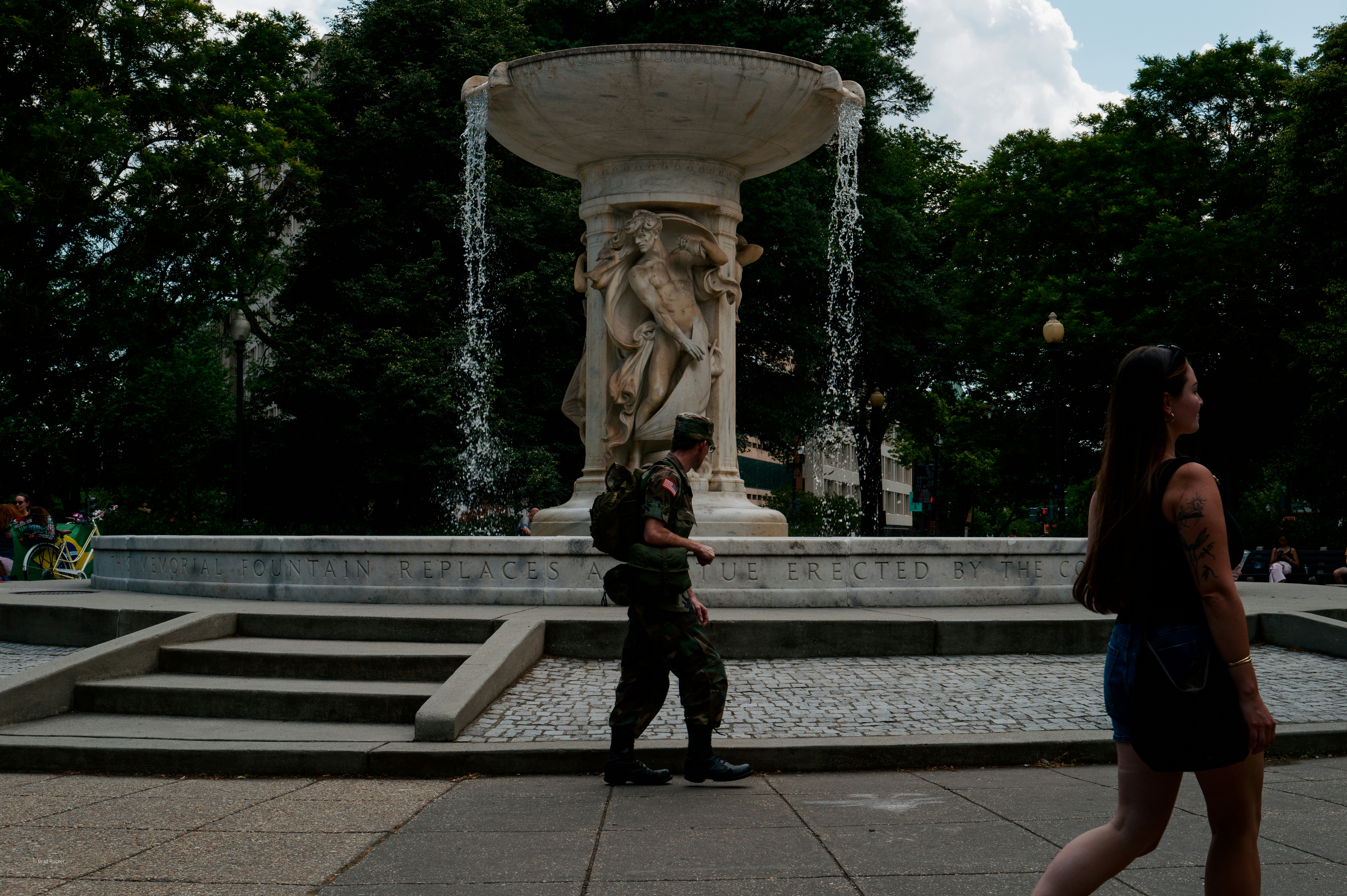 People walk past a large stone fountain in a park.