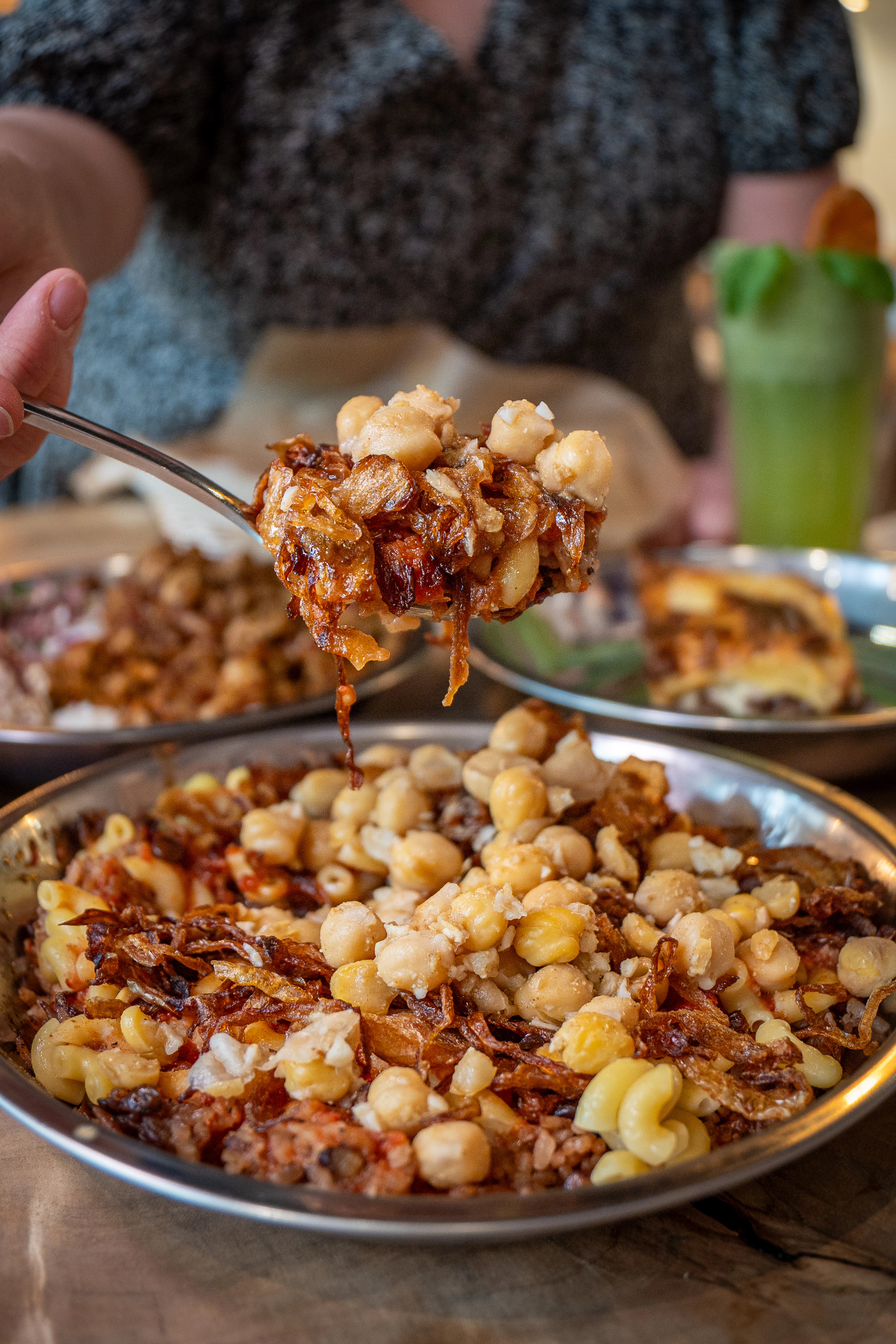 A metal dish of koshari, Egypt's national comfort dish of macaroni, lentils, rice and chickpeas under tomato sauce and crisp fried onions, with a spoonful lifted from it.