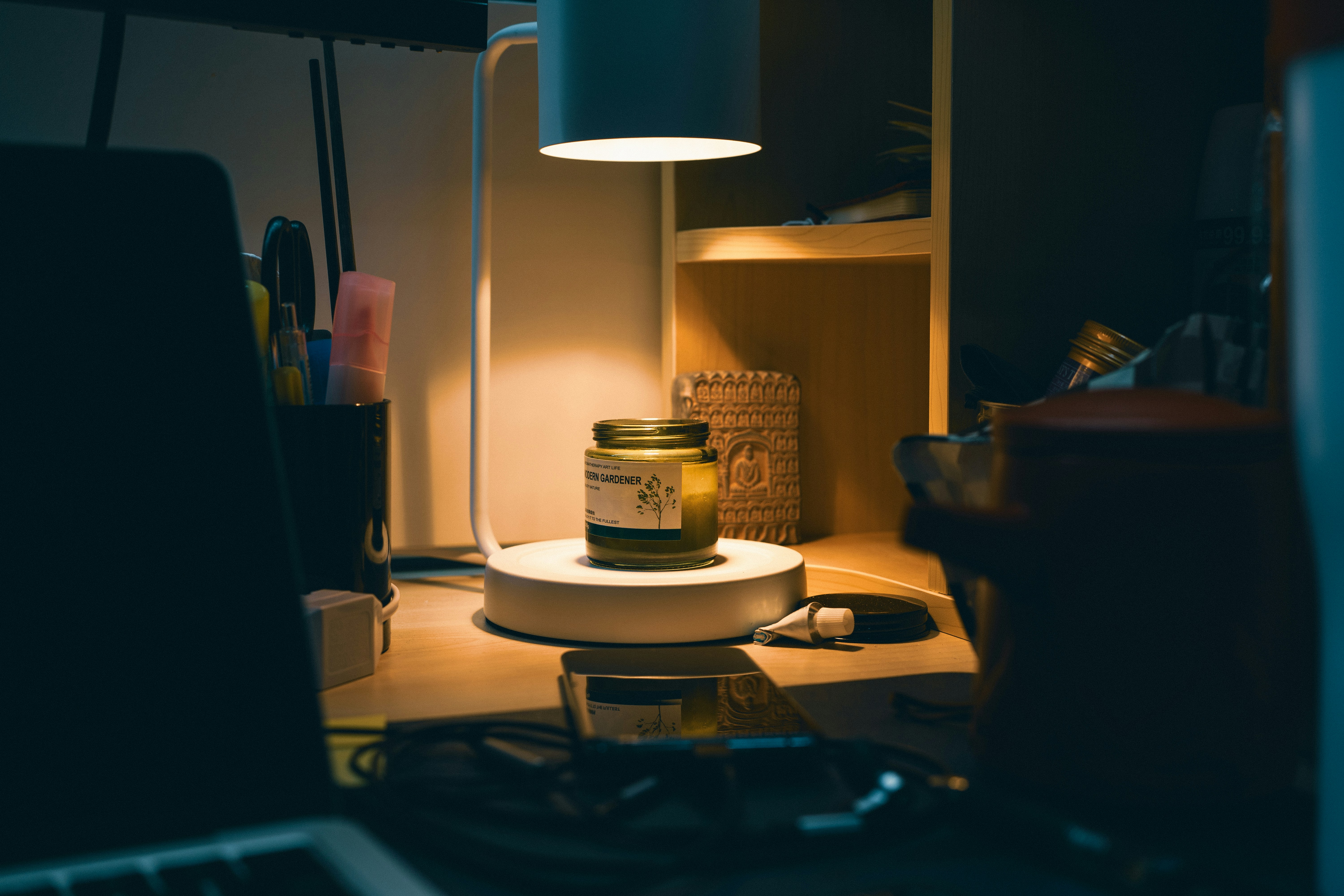 Desk lamp illuminates candle jar on a dimly lit workspace.
