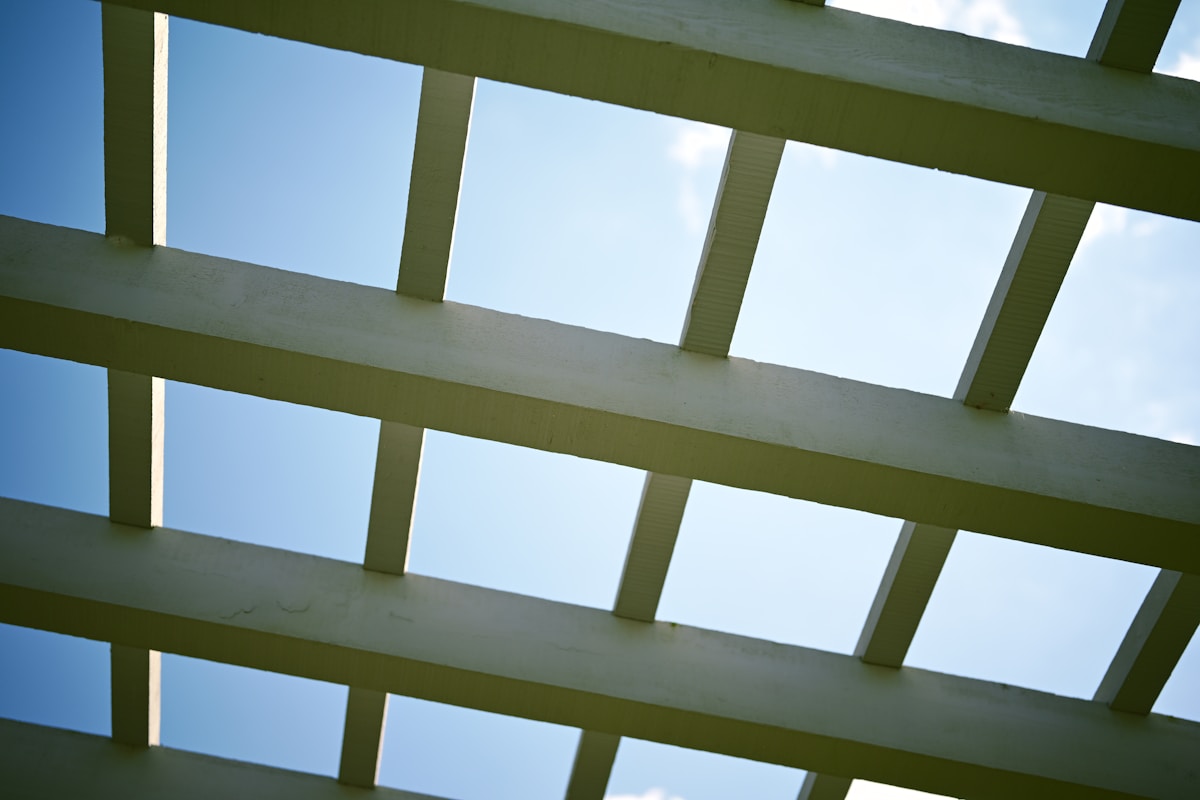 Looking up through a white wooden lattice pergola roof structure against a bright blue sky with scattered clouds