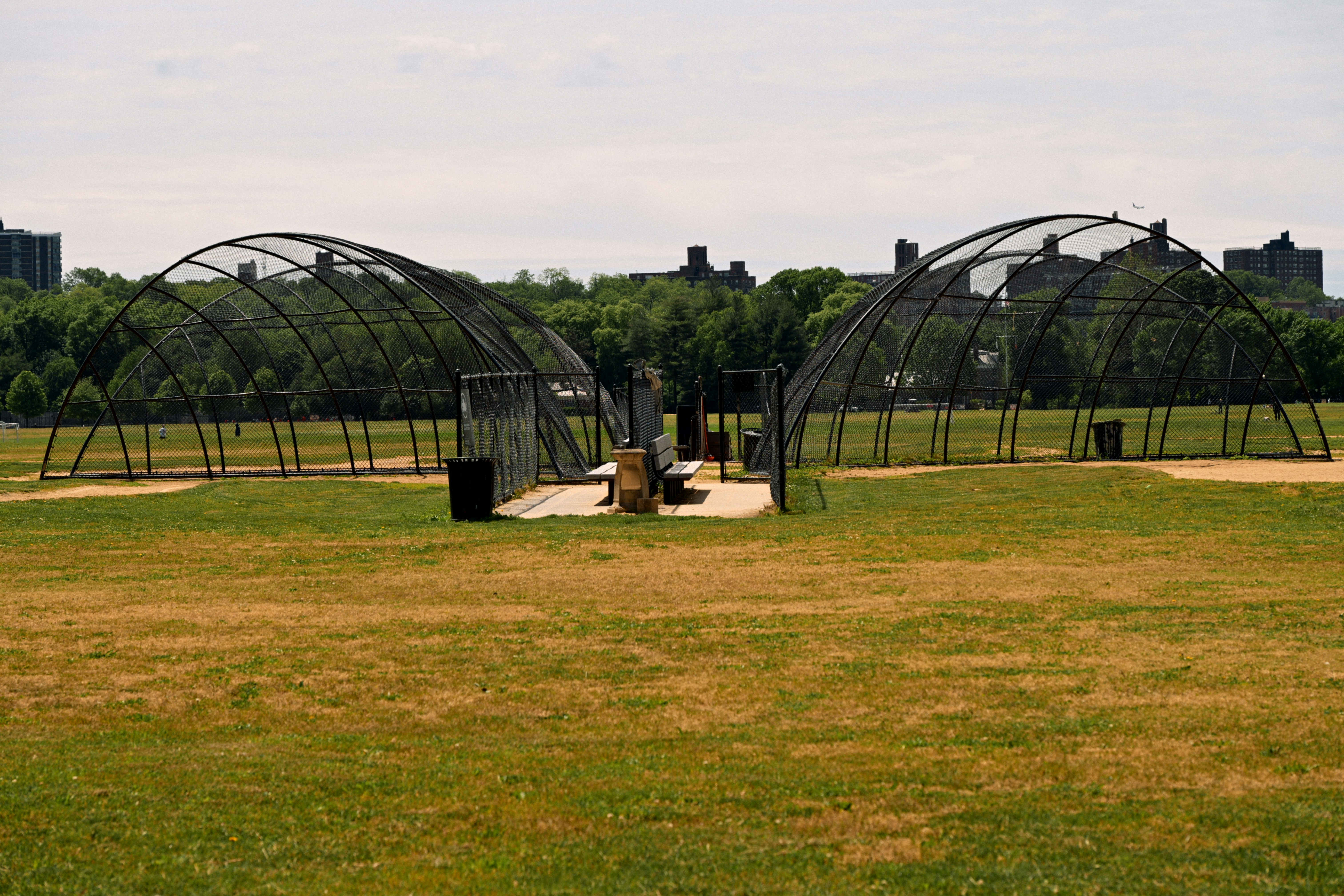 Baseball field with two batting cages and distant buildings.