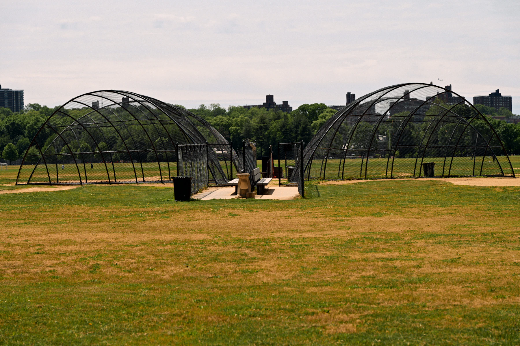 Two netted batting cages on a public field with trees behind