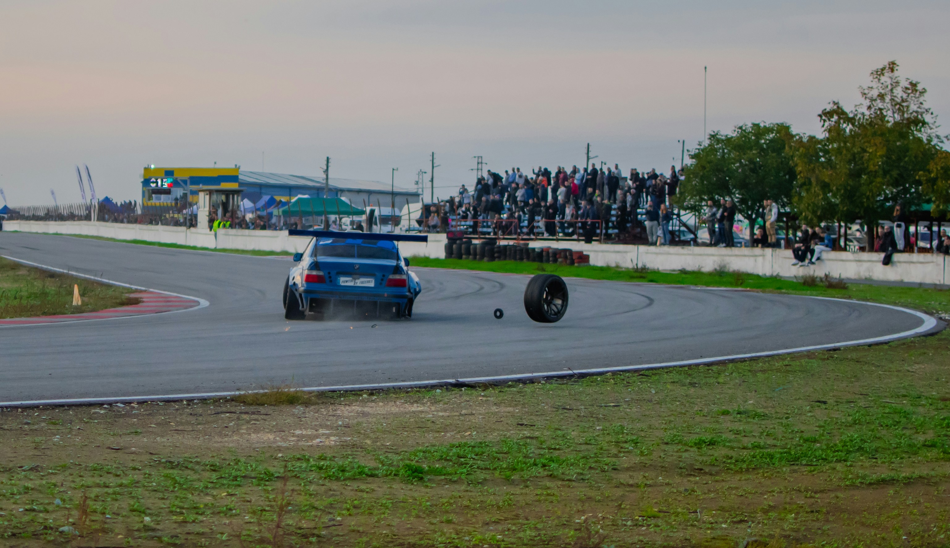 Blue race car on track, tire detached, crowd watching.