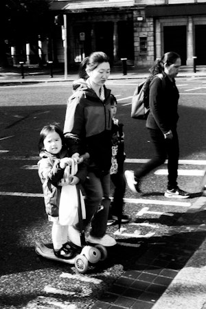A parent walking with two children, one riding a scooter, on a neighborhood sidewalk