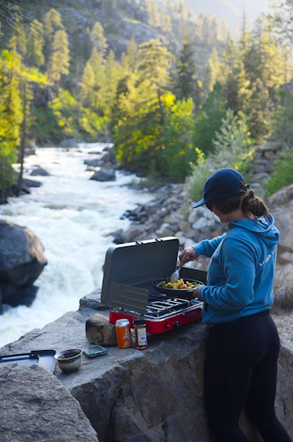 Woman cooking outdoors by a river with a portable stove.