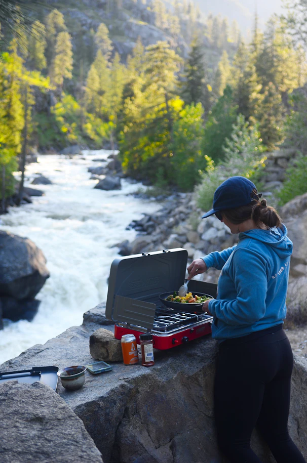 Woman cooking outdoors by a river with a portable stove.