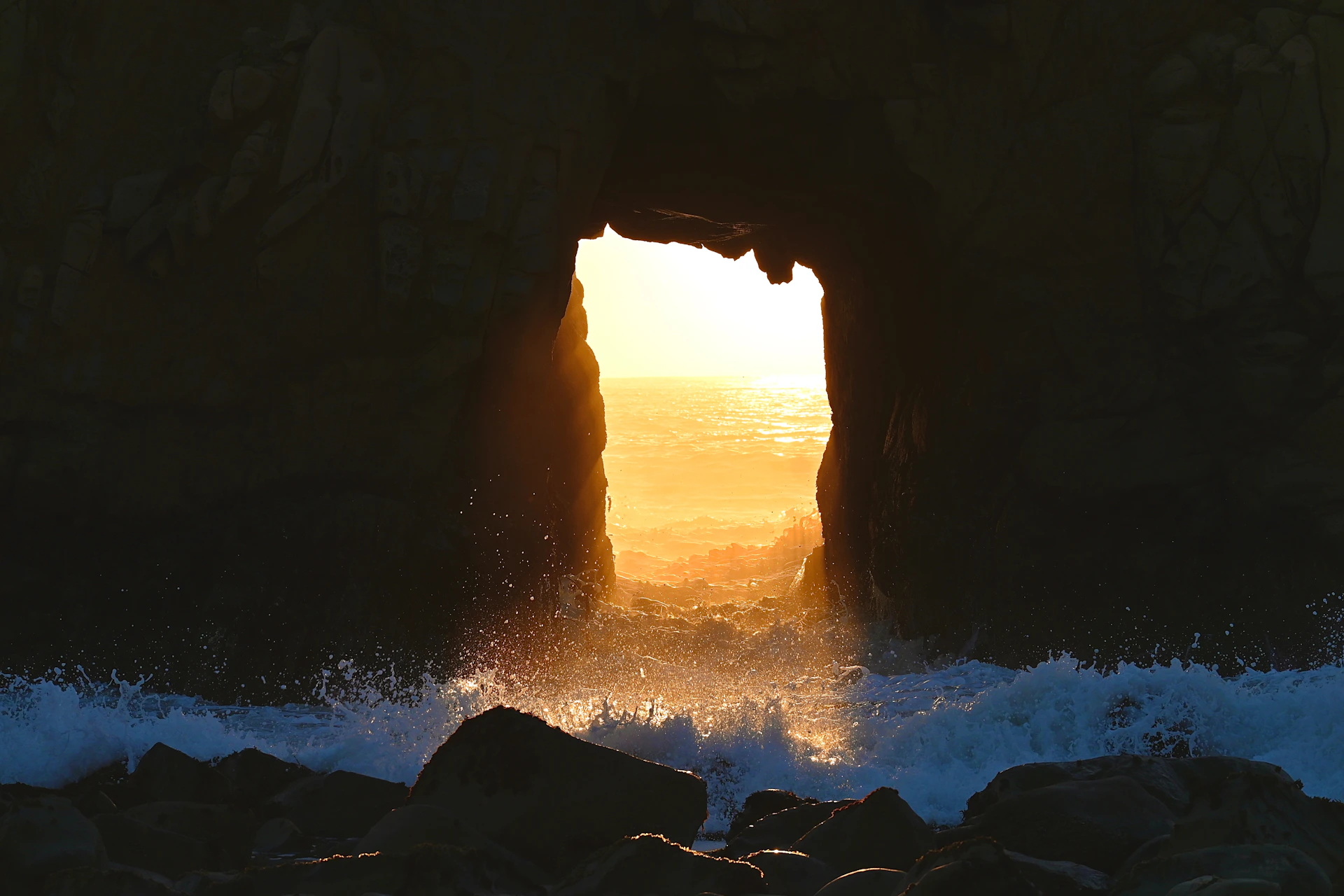 Ocean waves crash through a sunlit cave opening.