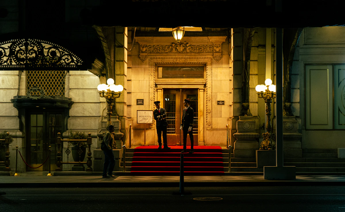 The lit entrance of a grand house, two doormen on duty on the red carpet