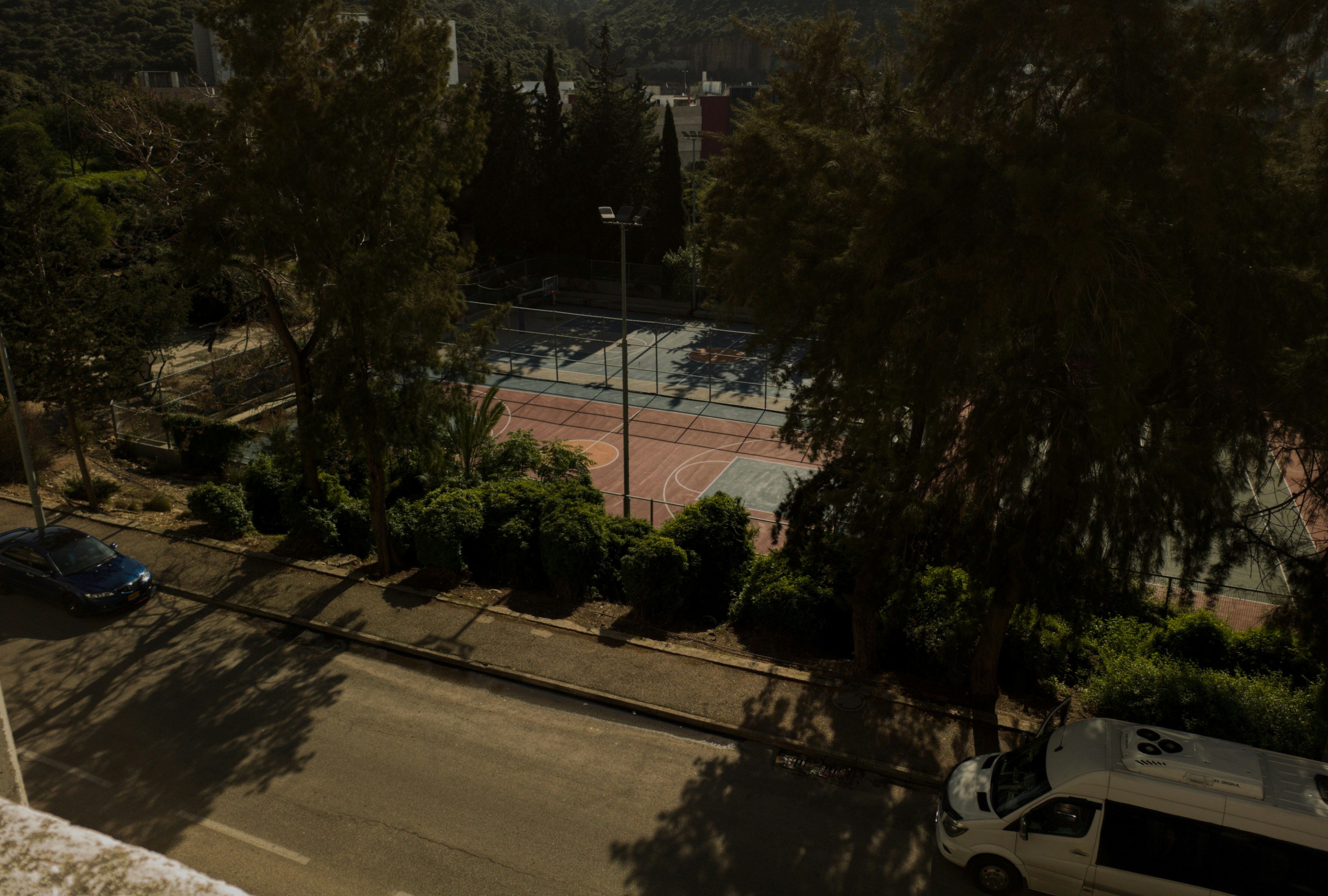 Un court multisports, une route avec des voitures et des arbres au soleil.