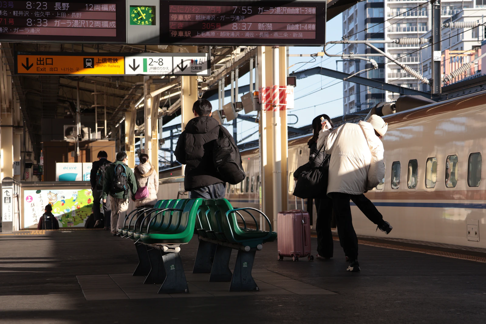 Photograph of travellers stand beside a high-speed train on a platform, with a pink wheeled suitcase nearby.