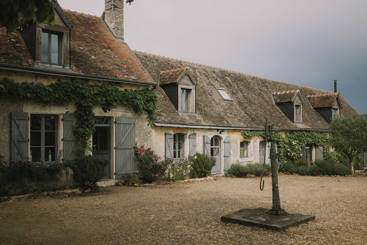 Une longère en pierre aux volets gris pâle, couverte de vigne et bordée de rosiers, devant une cour en gravier où se dresse une ancienne pompe à eau