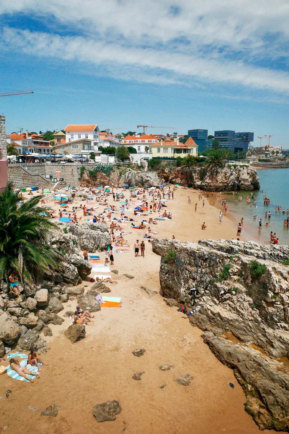 A sunny Atlantic beach below a Portuguese coastal town, packed with swimmers and framed by rocks