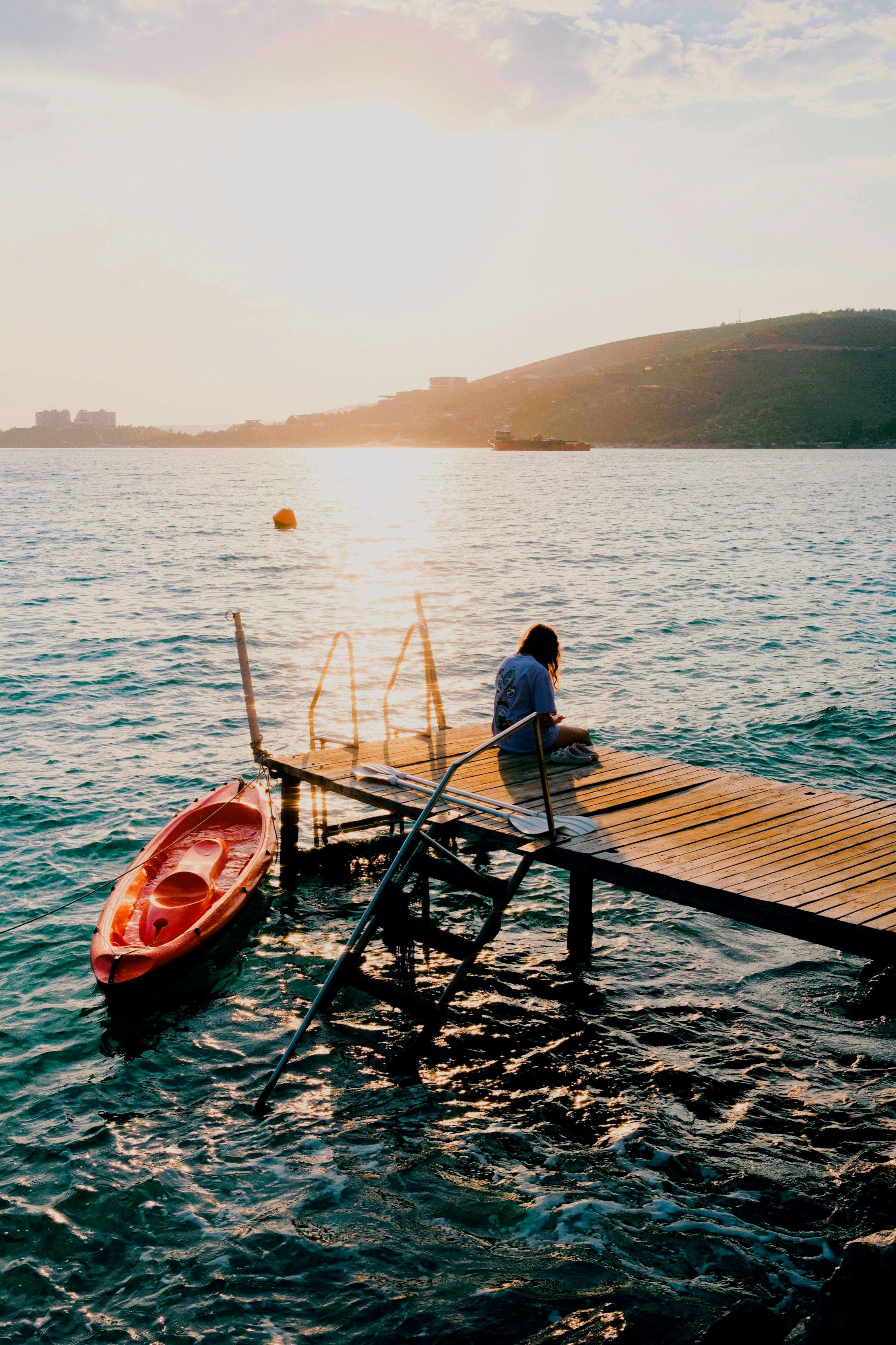 Person sits on pier with kayak at sunset.