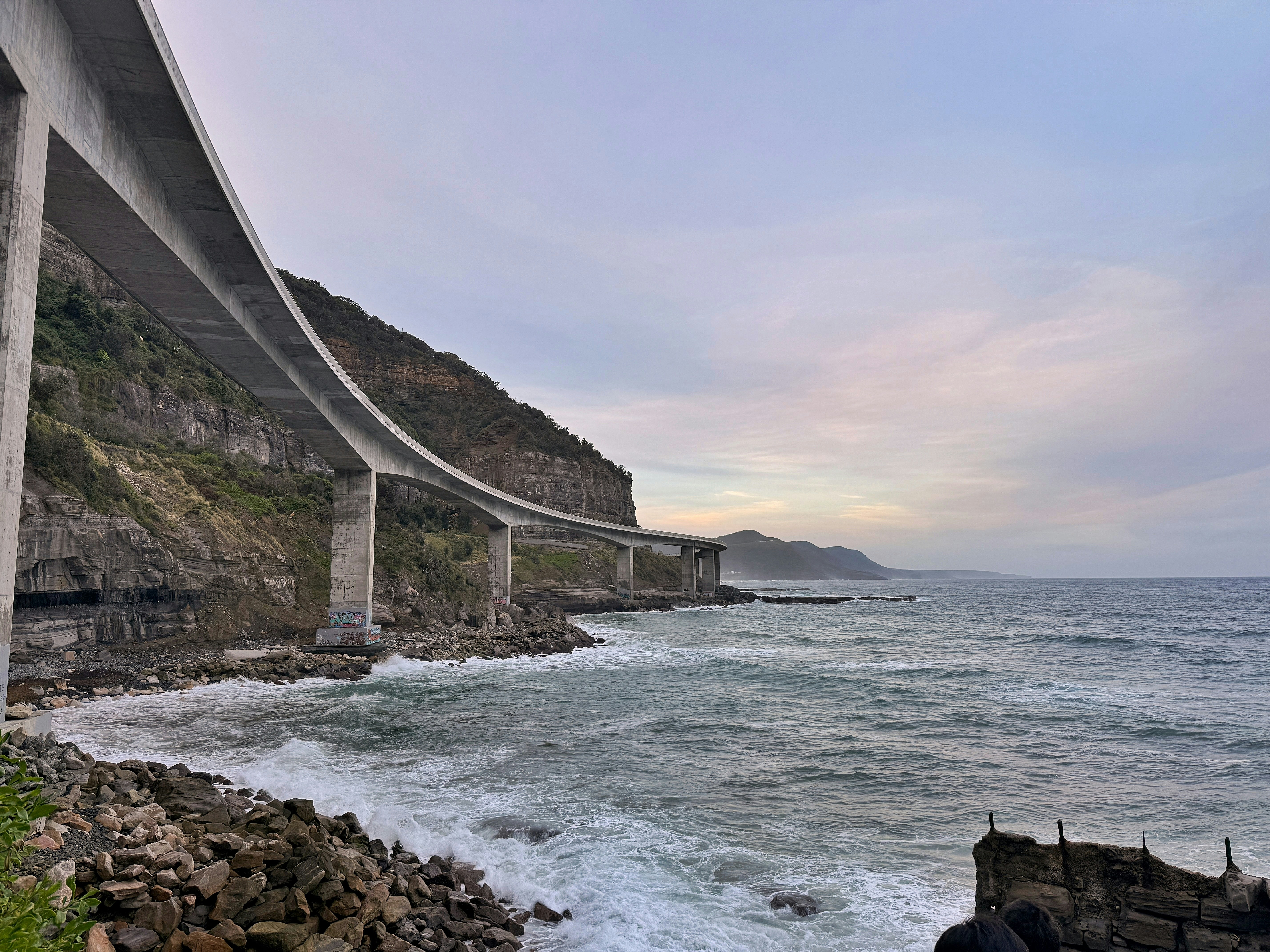 Coastal bridge extends along cliffs with ocean waves.