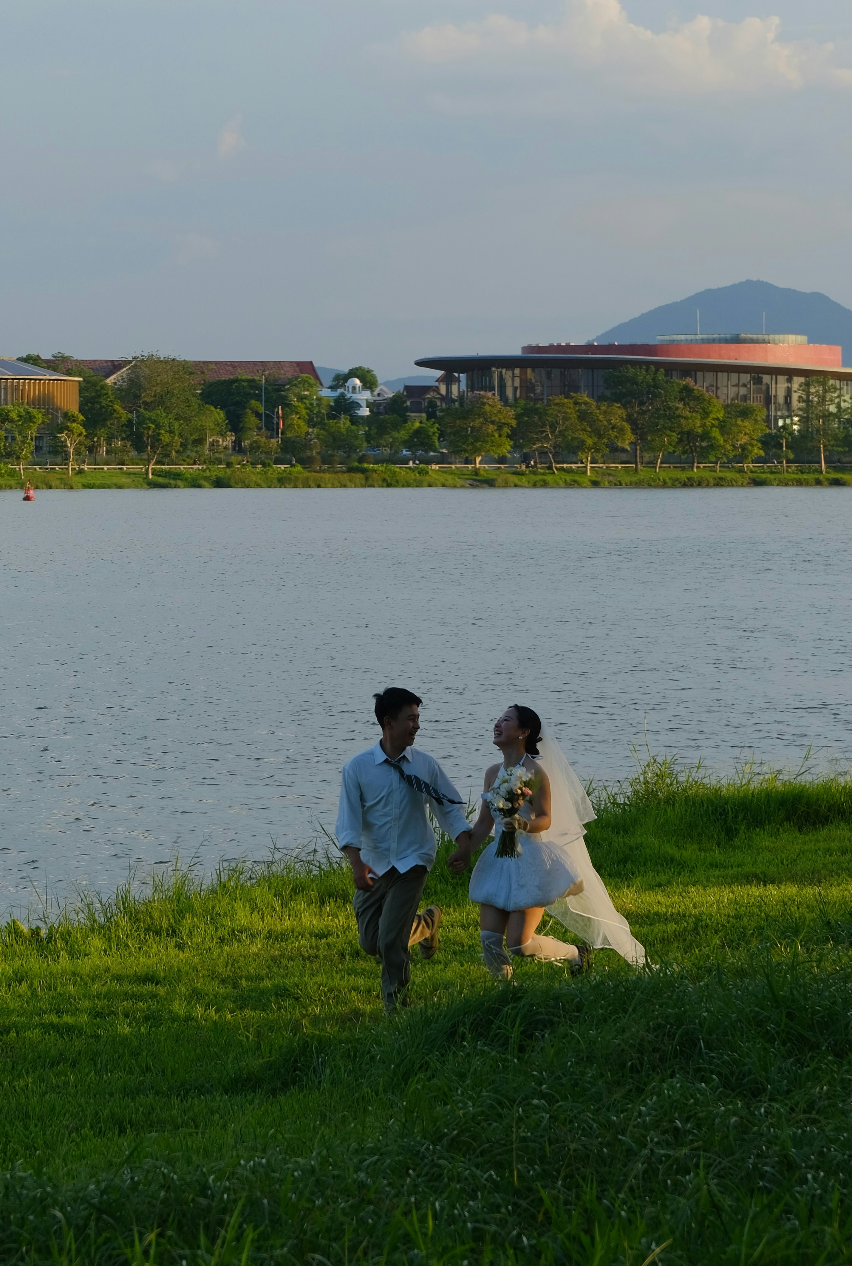 Los recién casados caminan de la mano por la orilla de un lago cubierto de hierba.