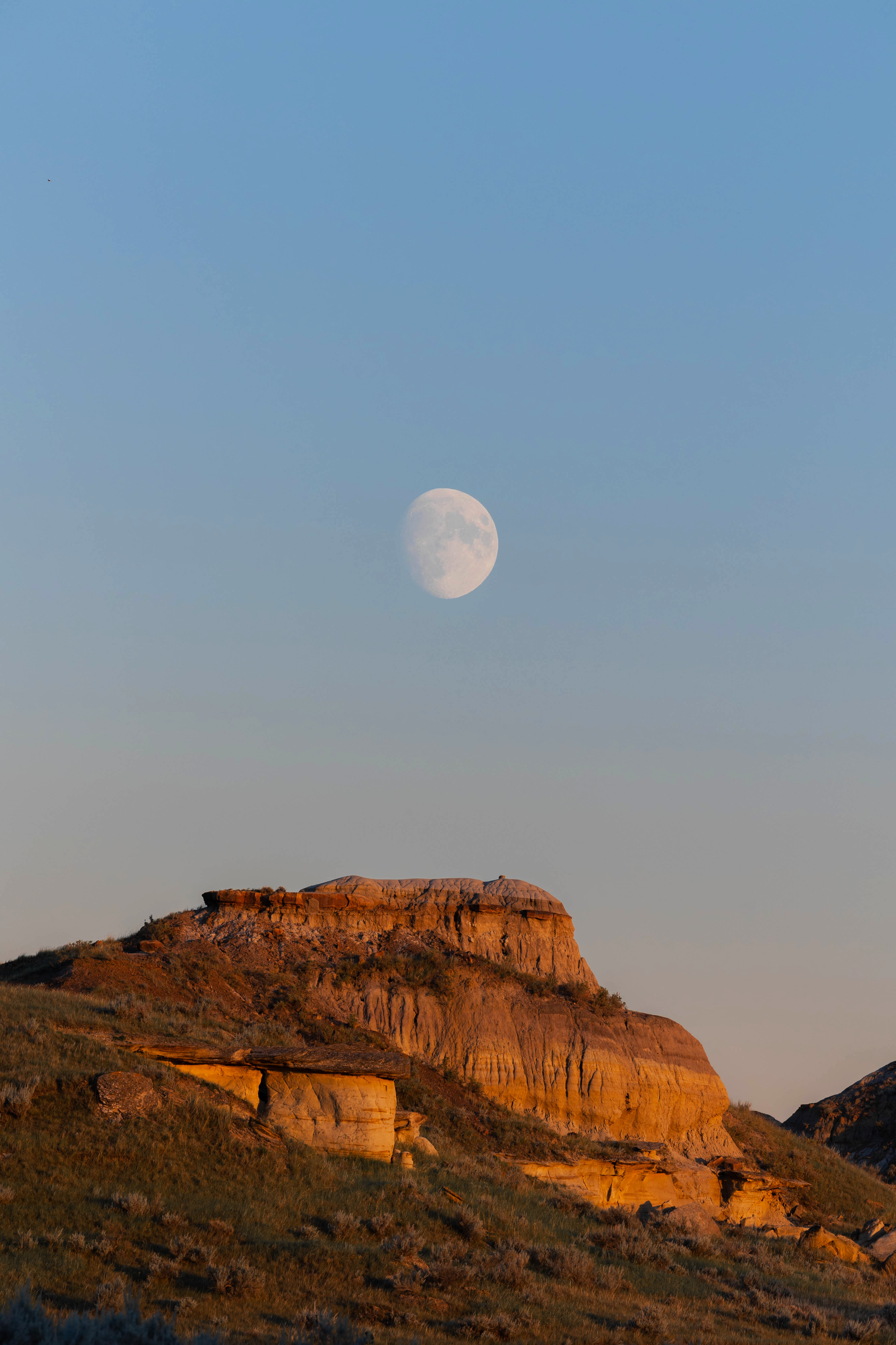 A bright moon hangs over a sunlit, rugged landscape.
