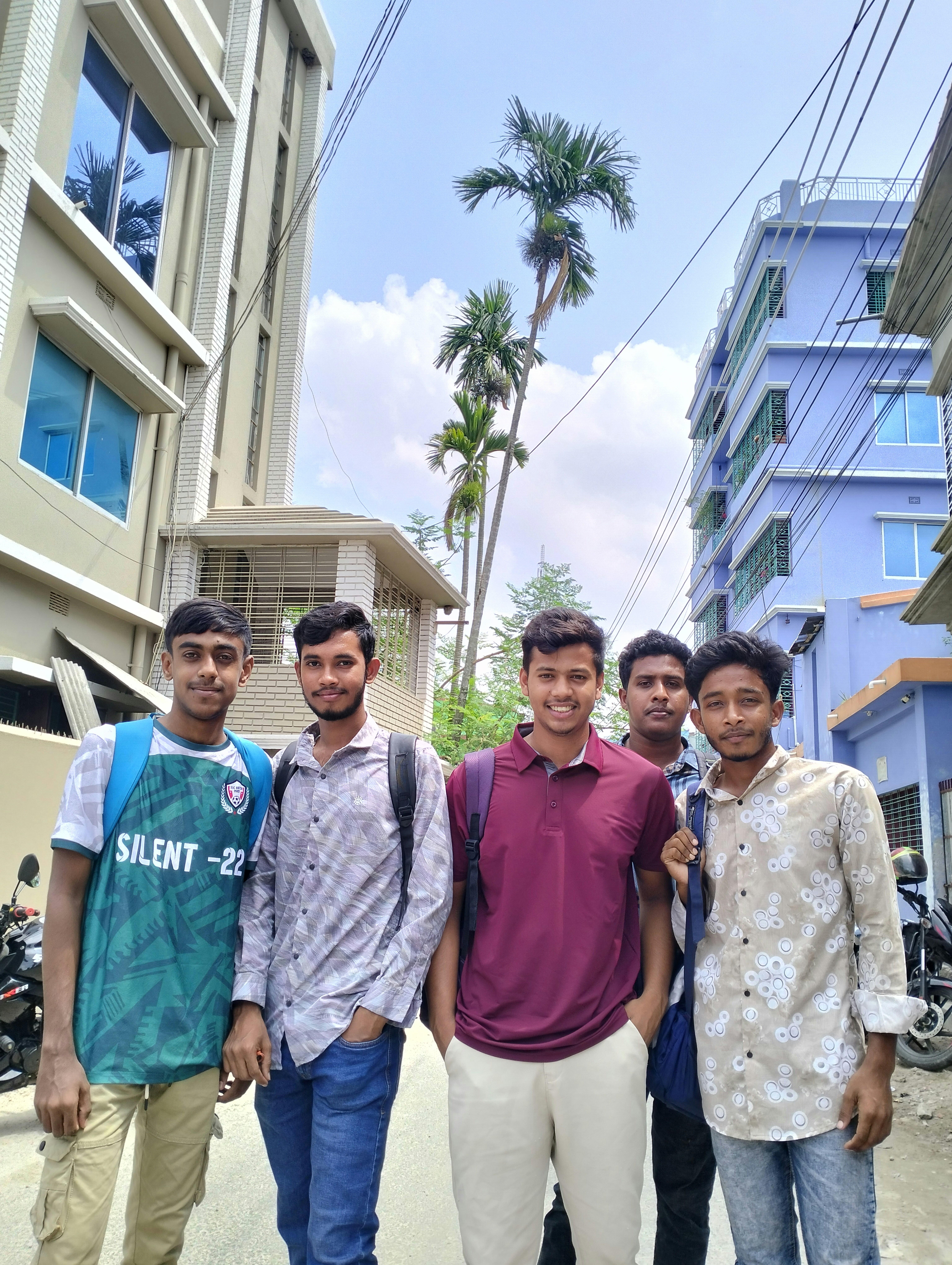 Five young men standing outdoors with buildings and trees.