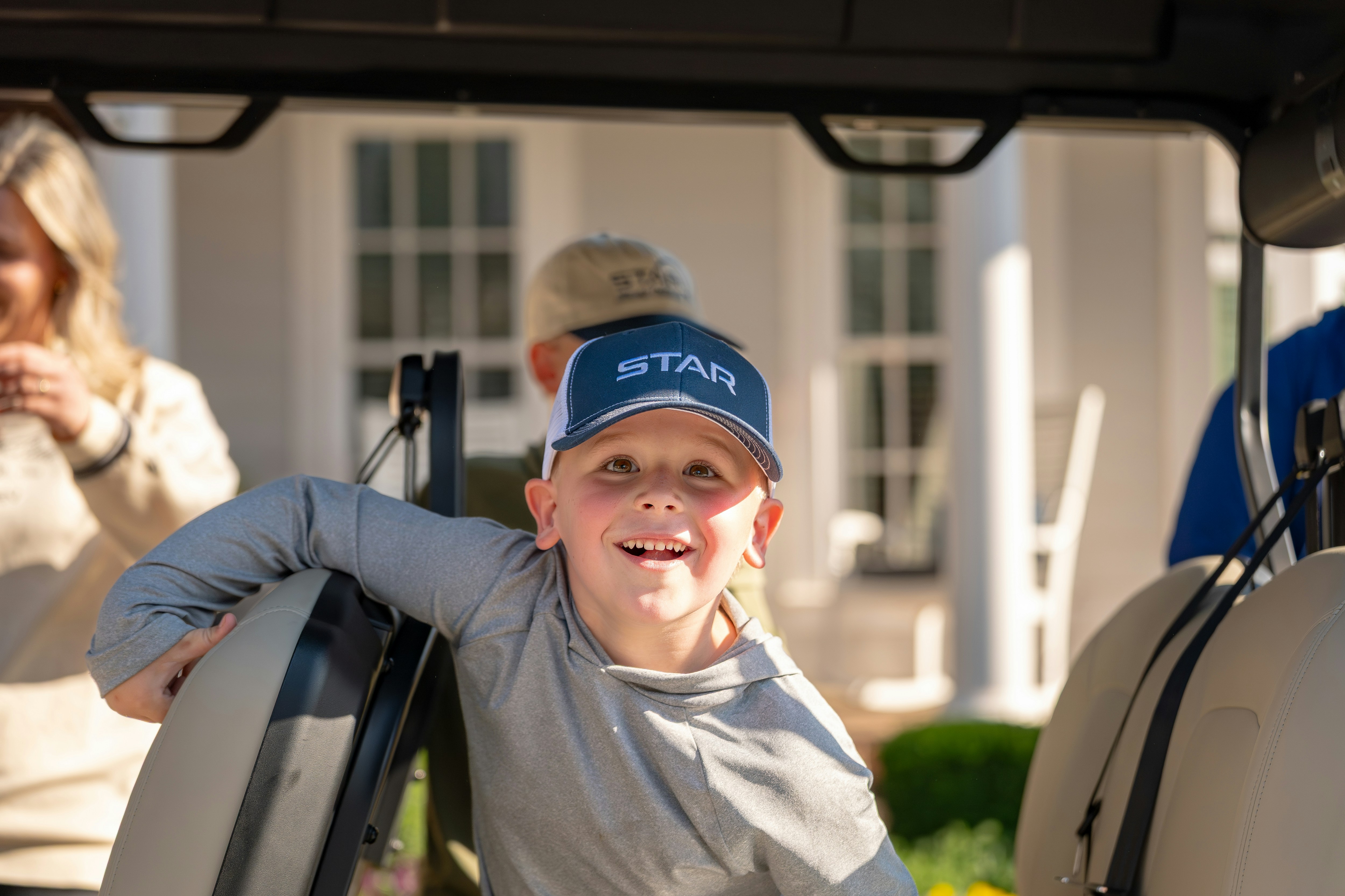 Smiling child sits in a golf cart with another person behind, sunny porch in the background; candid portrait highlighting a playful moment.
